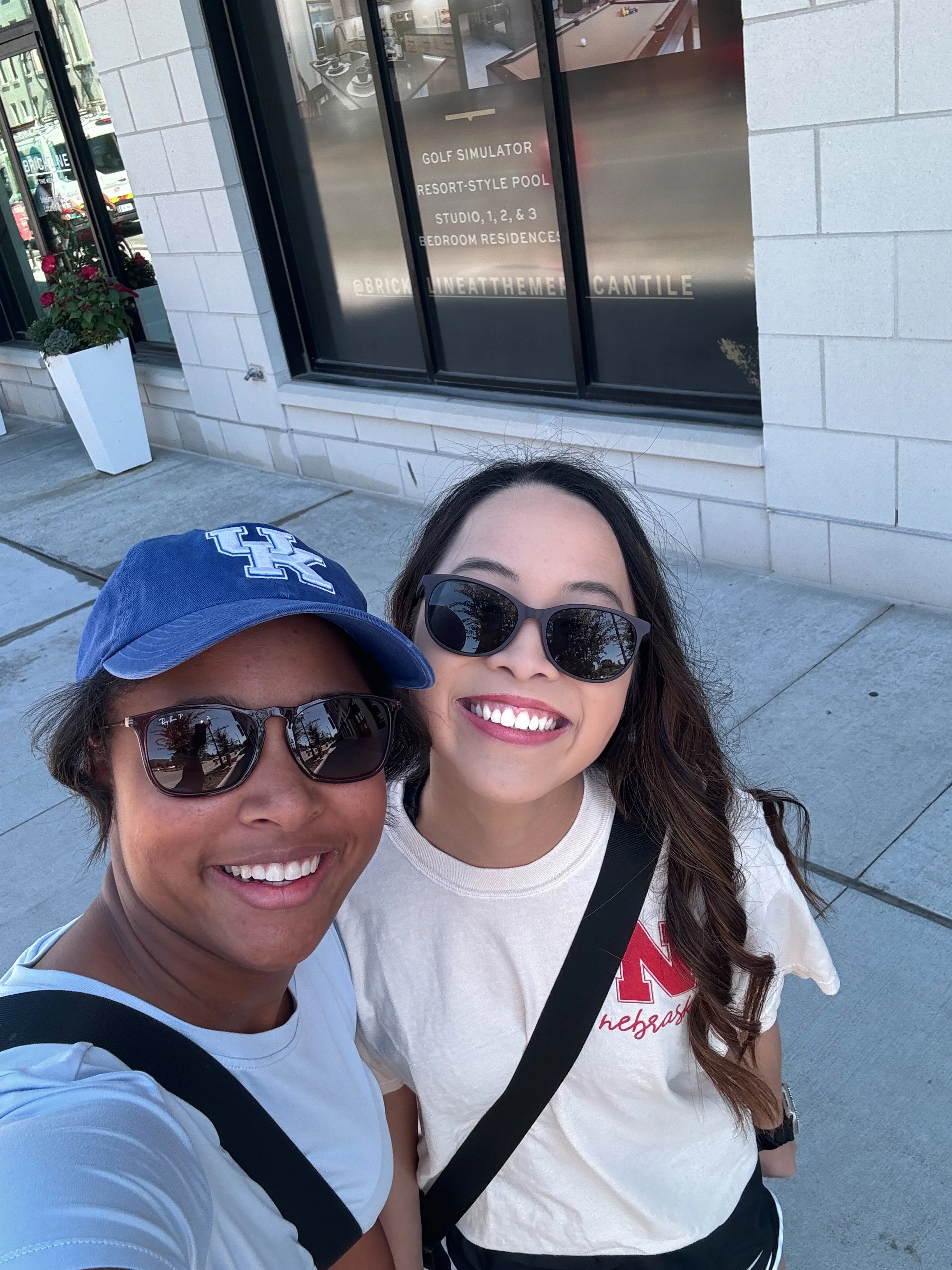 Two smiling women wearing sunglasses taking a selfie outside a building with large windows. One woman is wearing a blue baseball cap with a white H and a white T-shirt, the other is wearing a beige T-shirt with red lettering and a black strap across