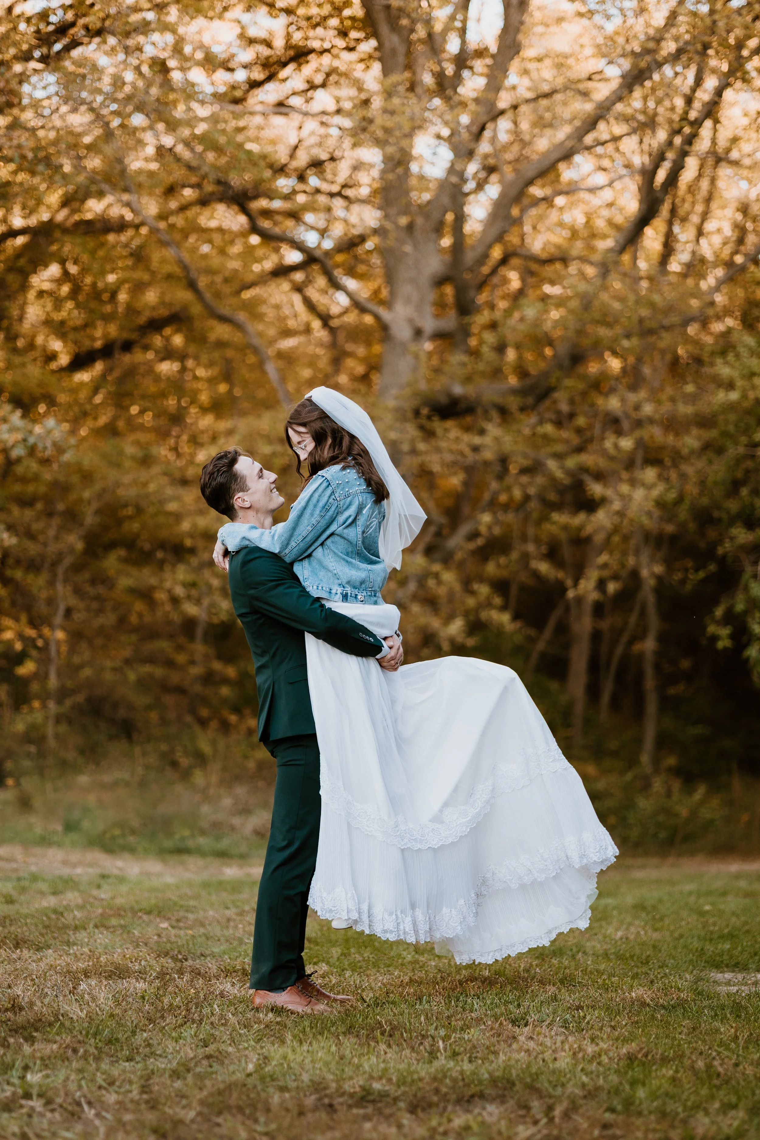 A groom lifting a bride in an outdoor setting during autumn, with trees and golden leaves in the background.