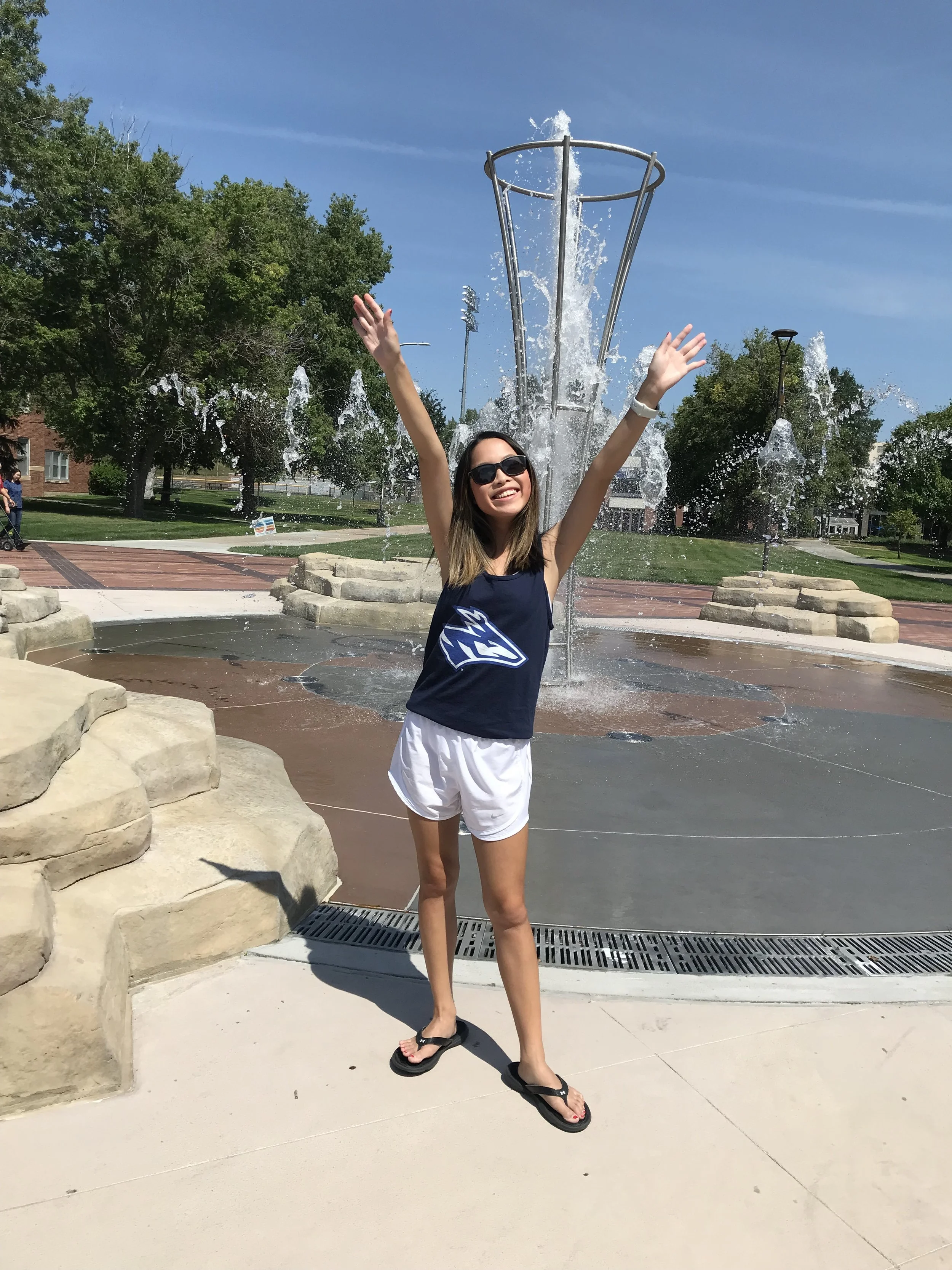 A woman in sunglasses, a navy tank top, and white shorts standing in front of a water fountain on a bright, sunny day, smiling with her arms raised.