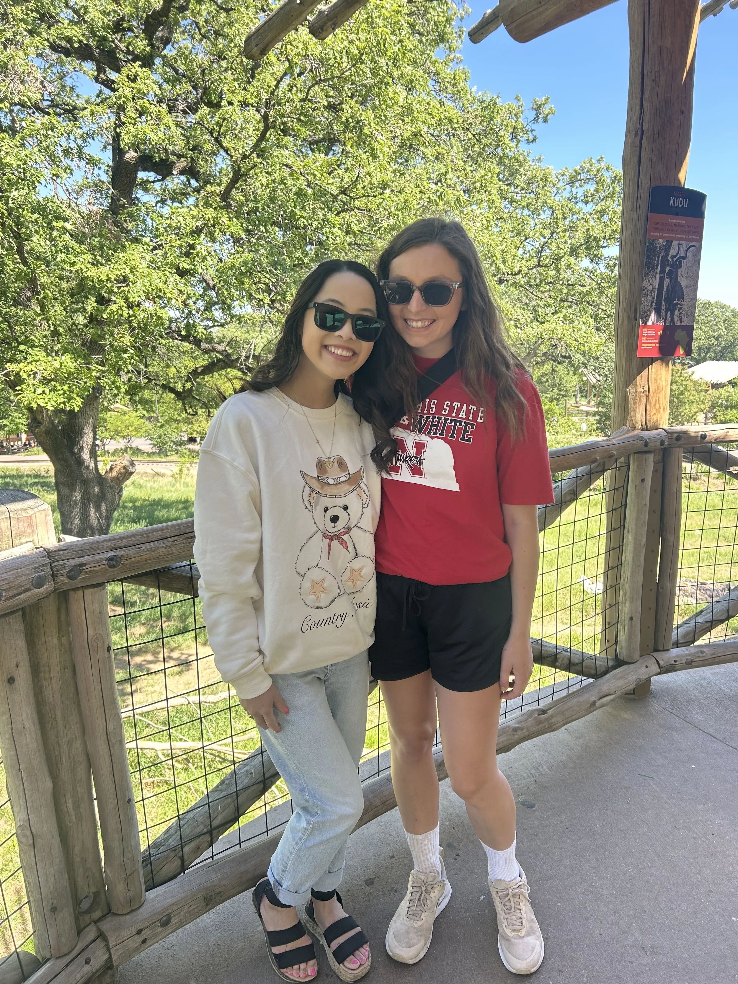 Two young women standing on a wooden walkway outdoors, smiling, wearing sunglasses, with green trees and blue sky in the background.