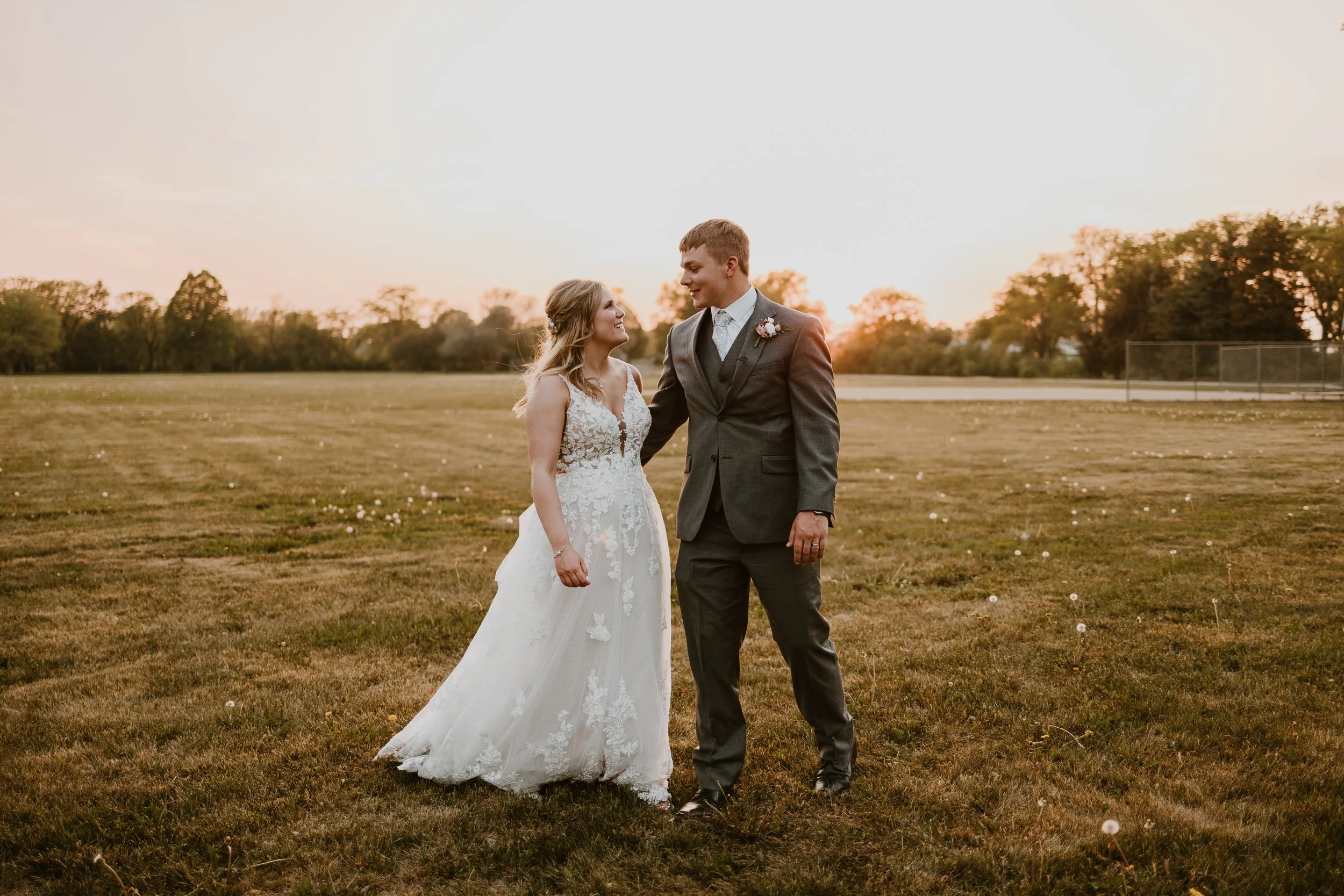 A newlywed couple stands close together on a field at sunset, with trees in the background. The bride wears a white lace wedding gown, and the groom wears a gray suit with a boutonniere. They look at each other lovingly.
