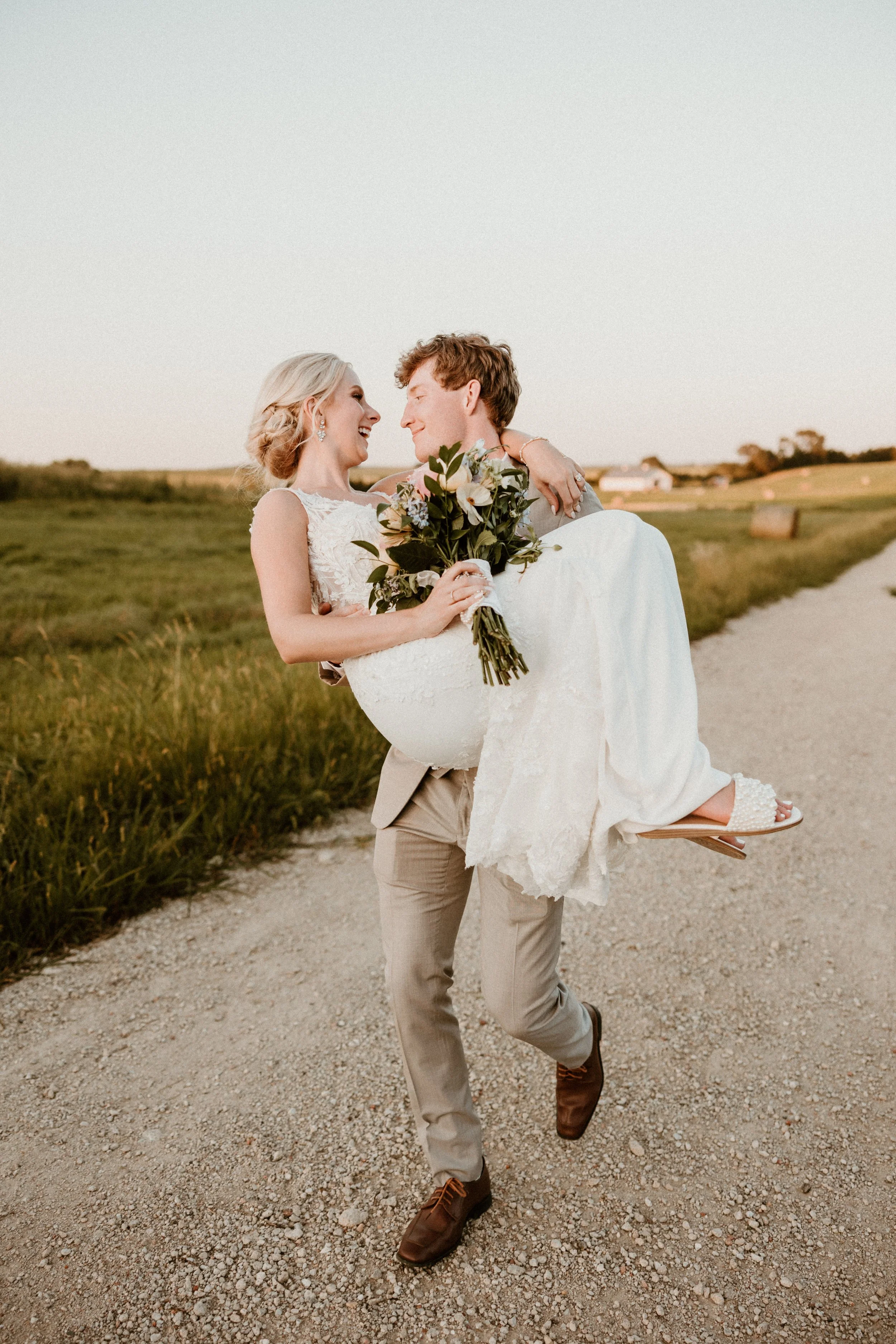 A man in beige trousers and a suit jacket carries a woman in a White lace wedding dress and white sandals on a gravel path in a rural setting with green fields and hay bales, holding a bouquet of flowers, as they smile at each other during sunset.