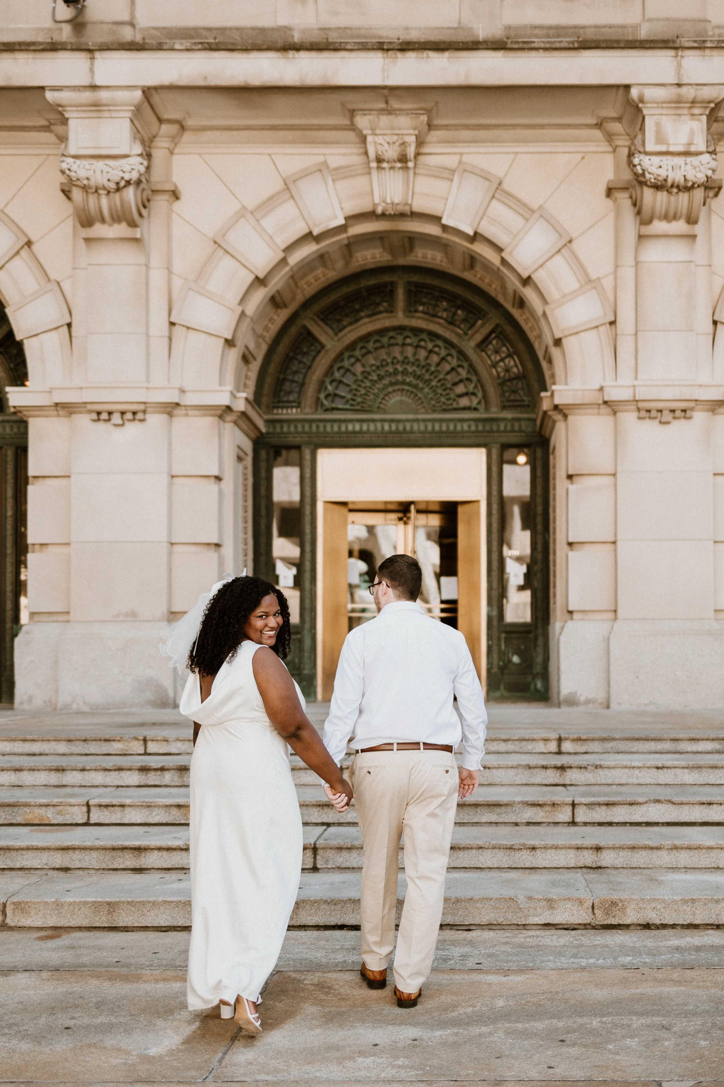 A couple holding hands and walking up the steps of a historic building, smiling at each other, with the woman wearing a white dress and veil.