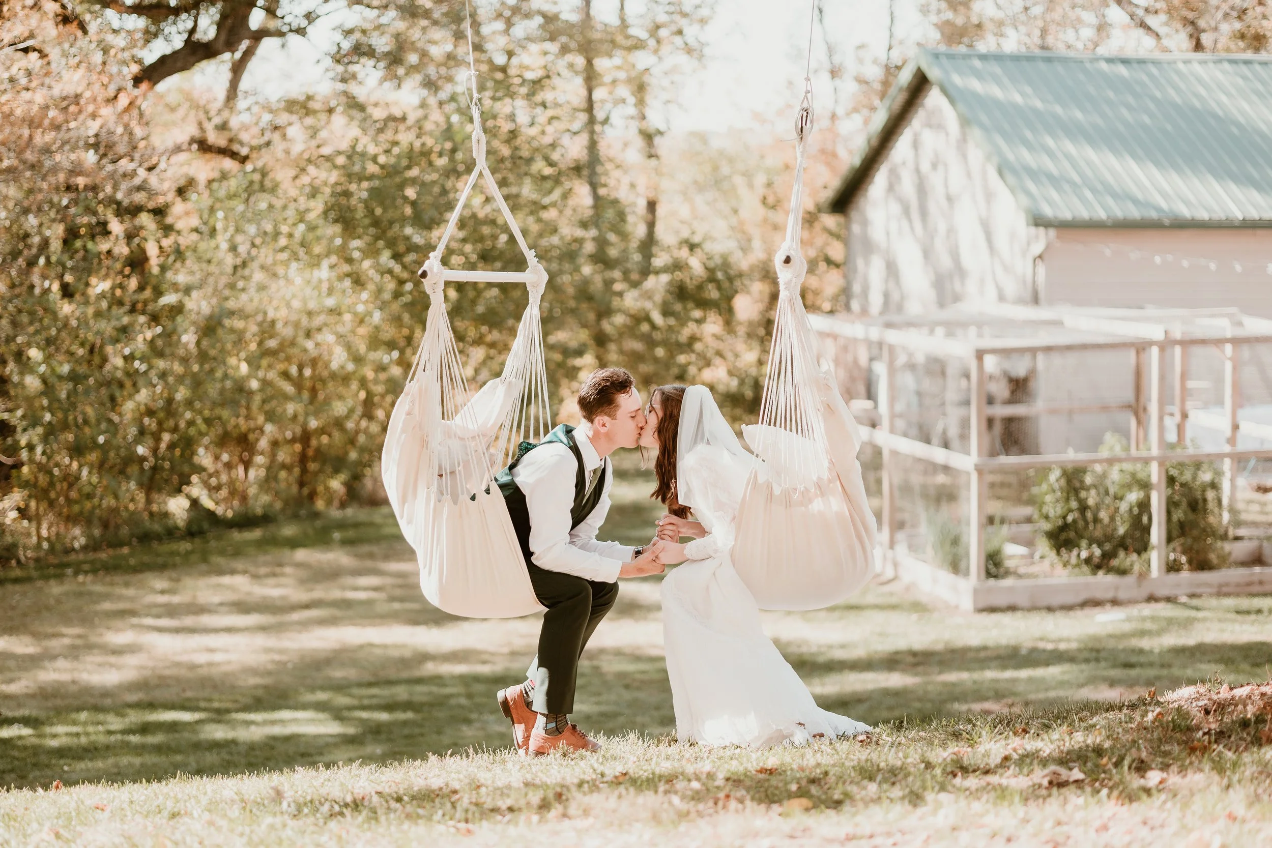 A bride and groom kiss while sitting on hanging fabric swings outdoors in a park or garden during daytime.