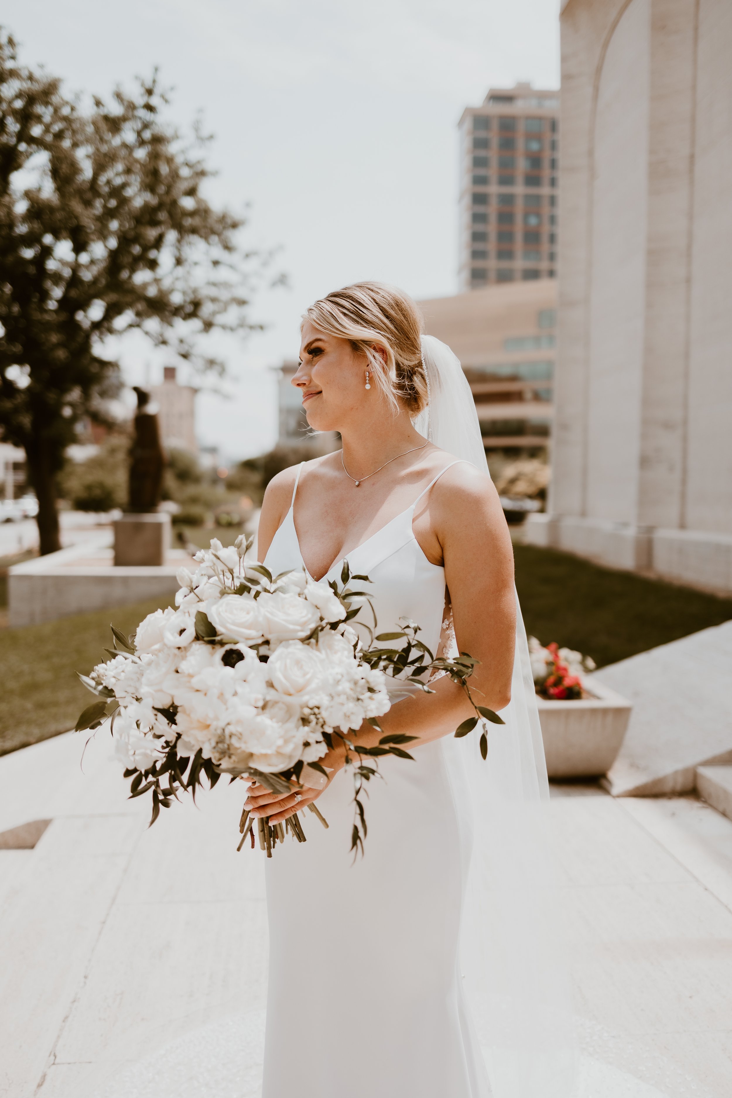 A bride in a white wedding dress holding a bouquet of white flowers outdoors in an urban setting.