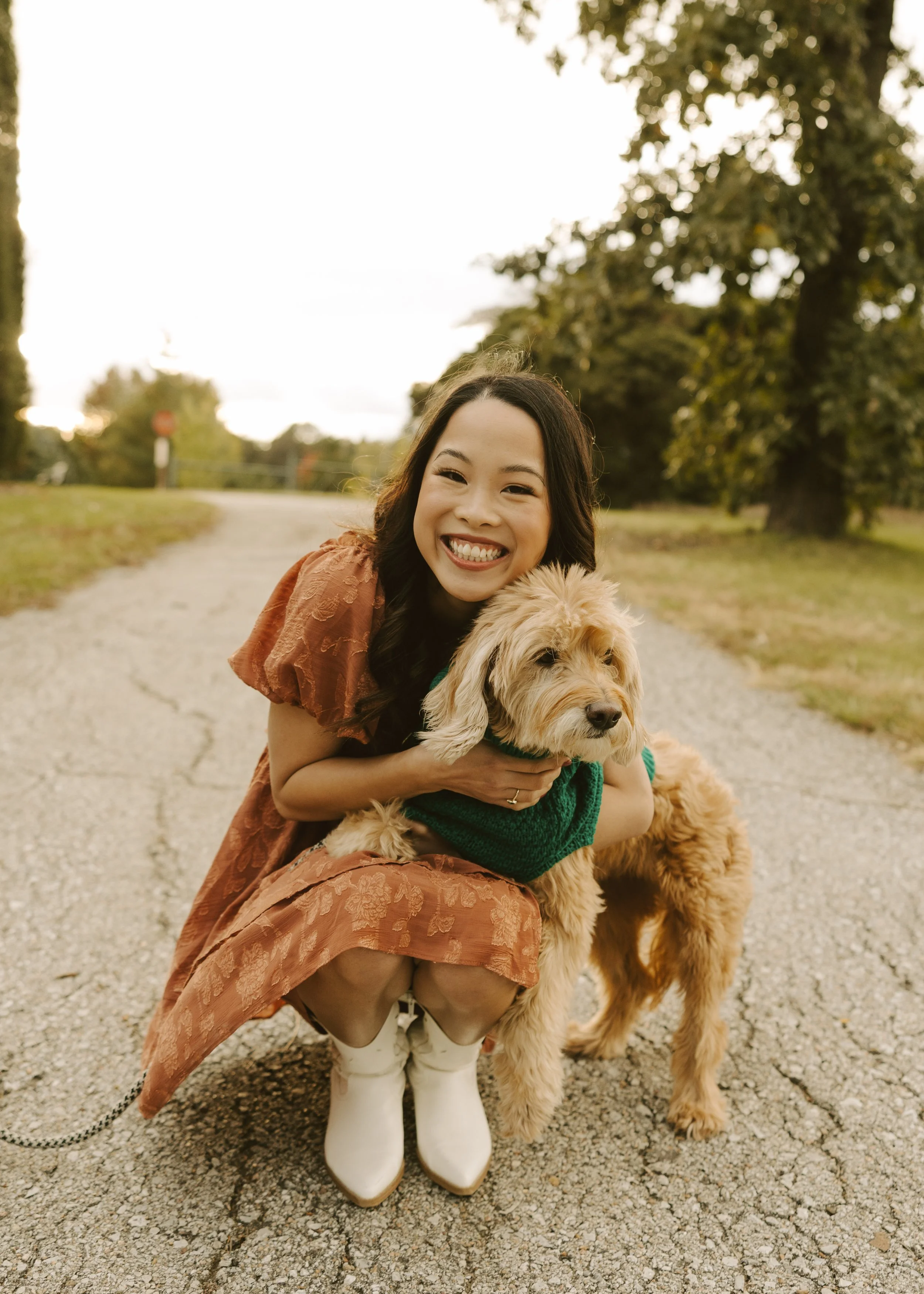 A smiling woman in an orange dress crouches outdoors on a gravel path, holding a fluffy golden-colored dog wearing a green sweater, with trees and cloudy sky in the background.