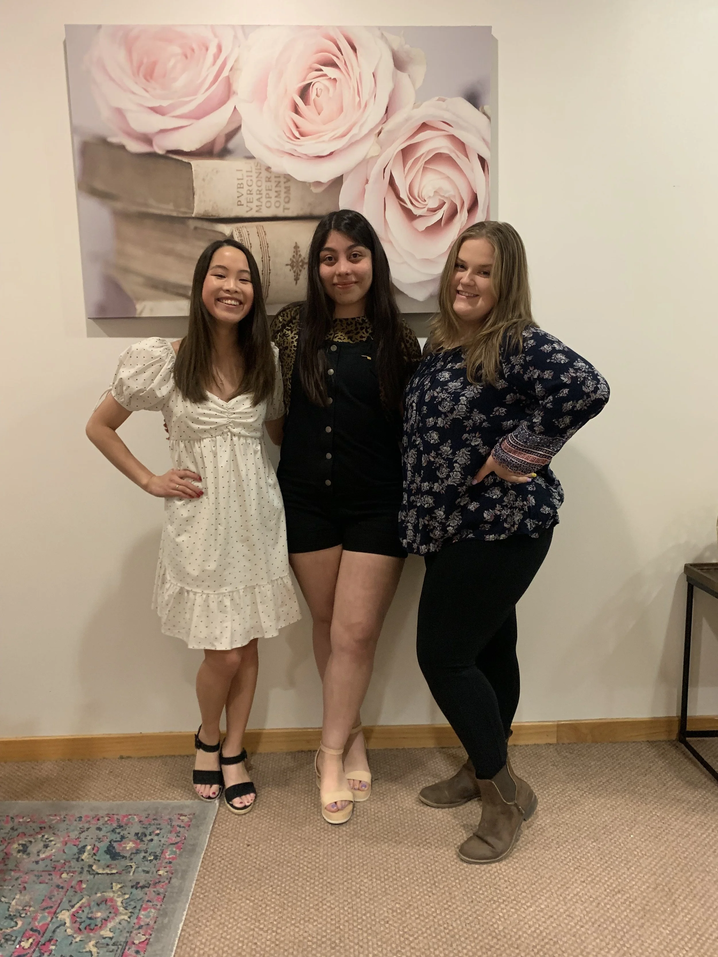 Three women standing in front of a large floral artwork, smiling at the camera. The woman on the left is wearing a white dress with polka dots, the woman in the center is wearing black shorts and a black top with leopard print details, and the woman