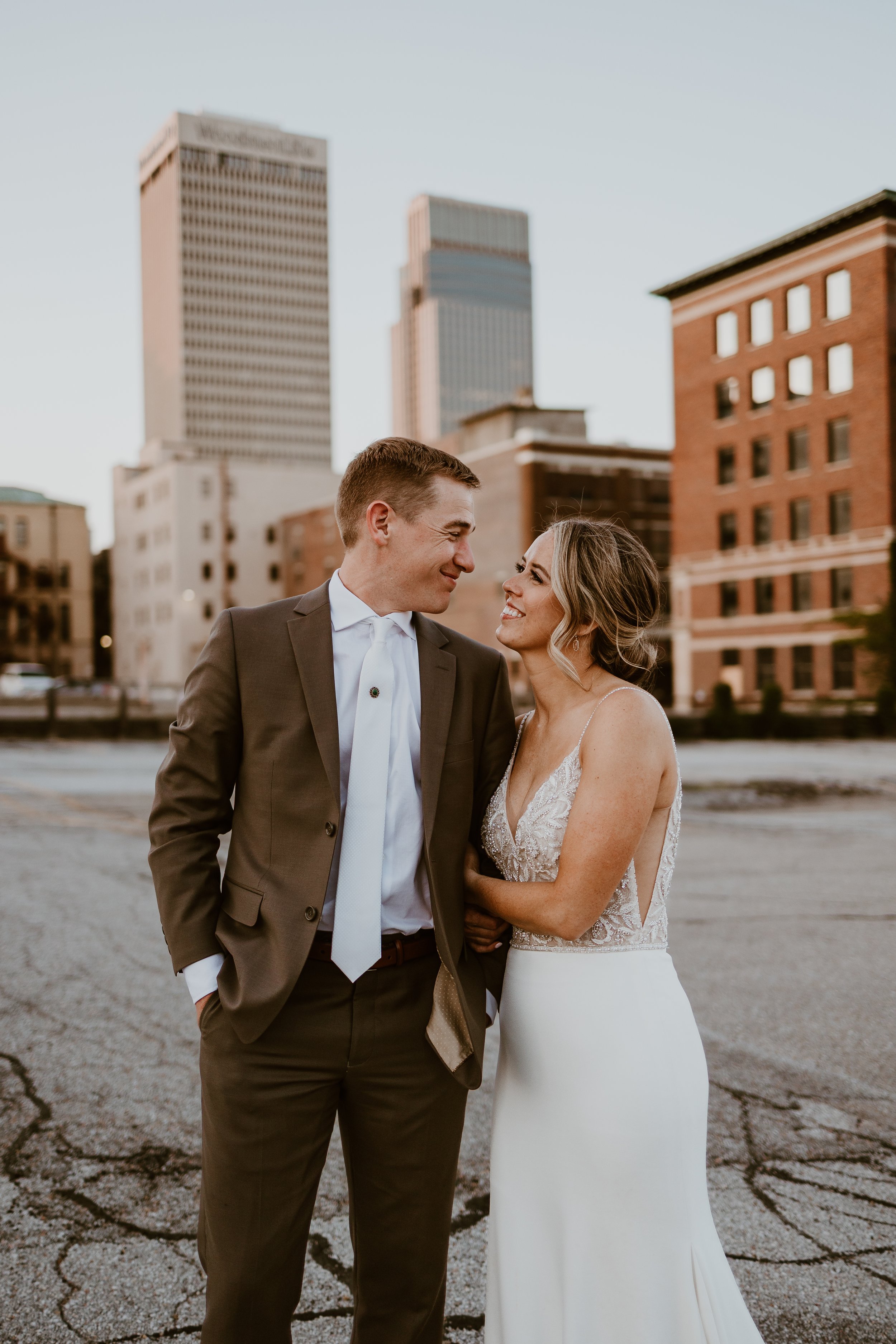 A newlywed couple smiling at each other outdoors with city buildings in the background during sunset.