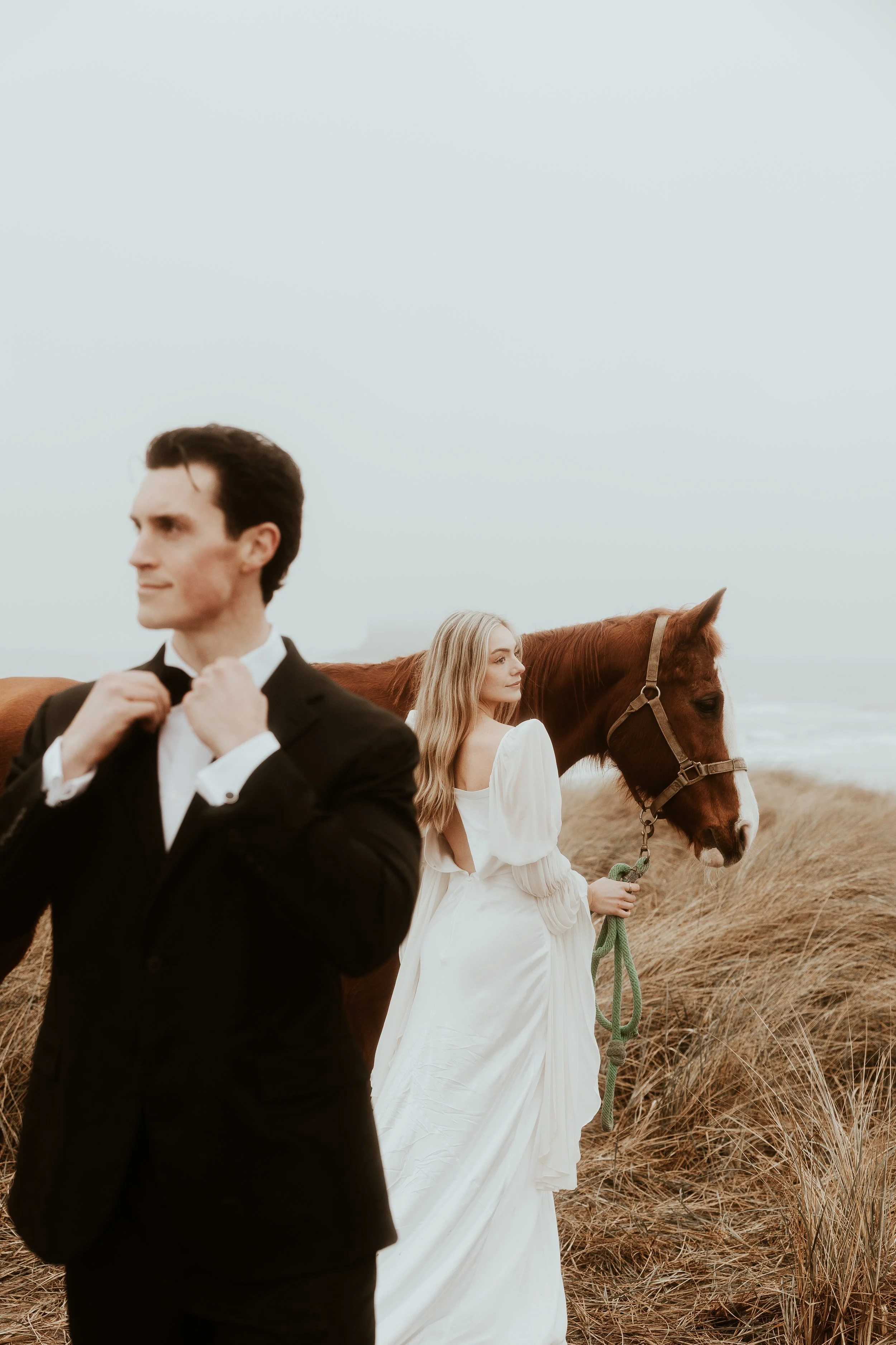 A man in a tuxedo adjusting his bow tie stands in the foreground, with a woman in a white dress holding a brown horse by the reins in a grassy field near the coast.