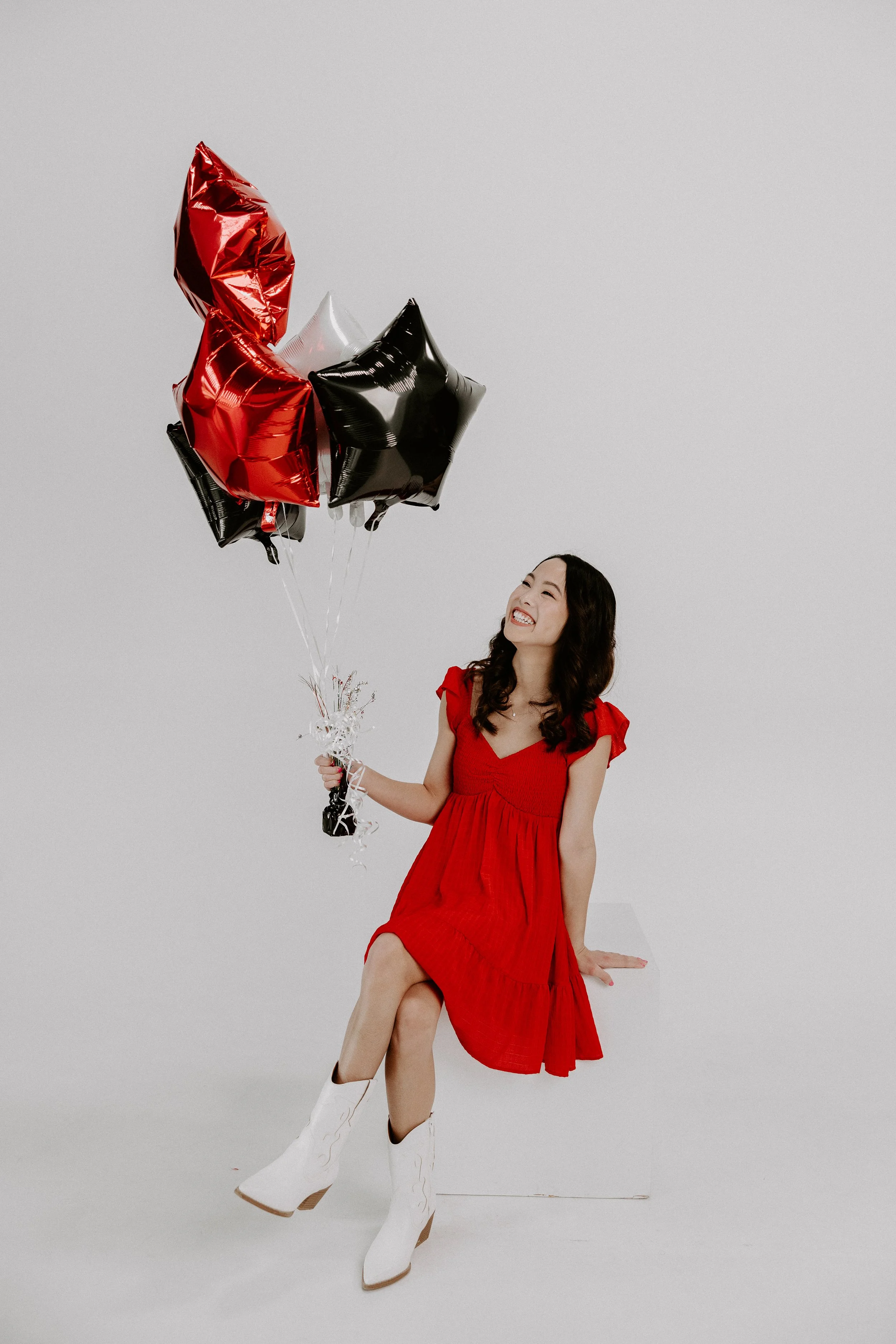 A young woman in a red dress and white cowboy boots sitting on a white box, holding a bunch of black, red, and white star-shaped balloons, smiling against a plain white background.