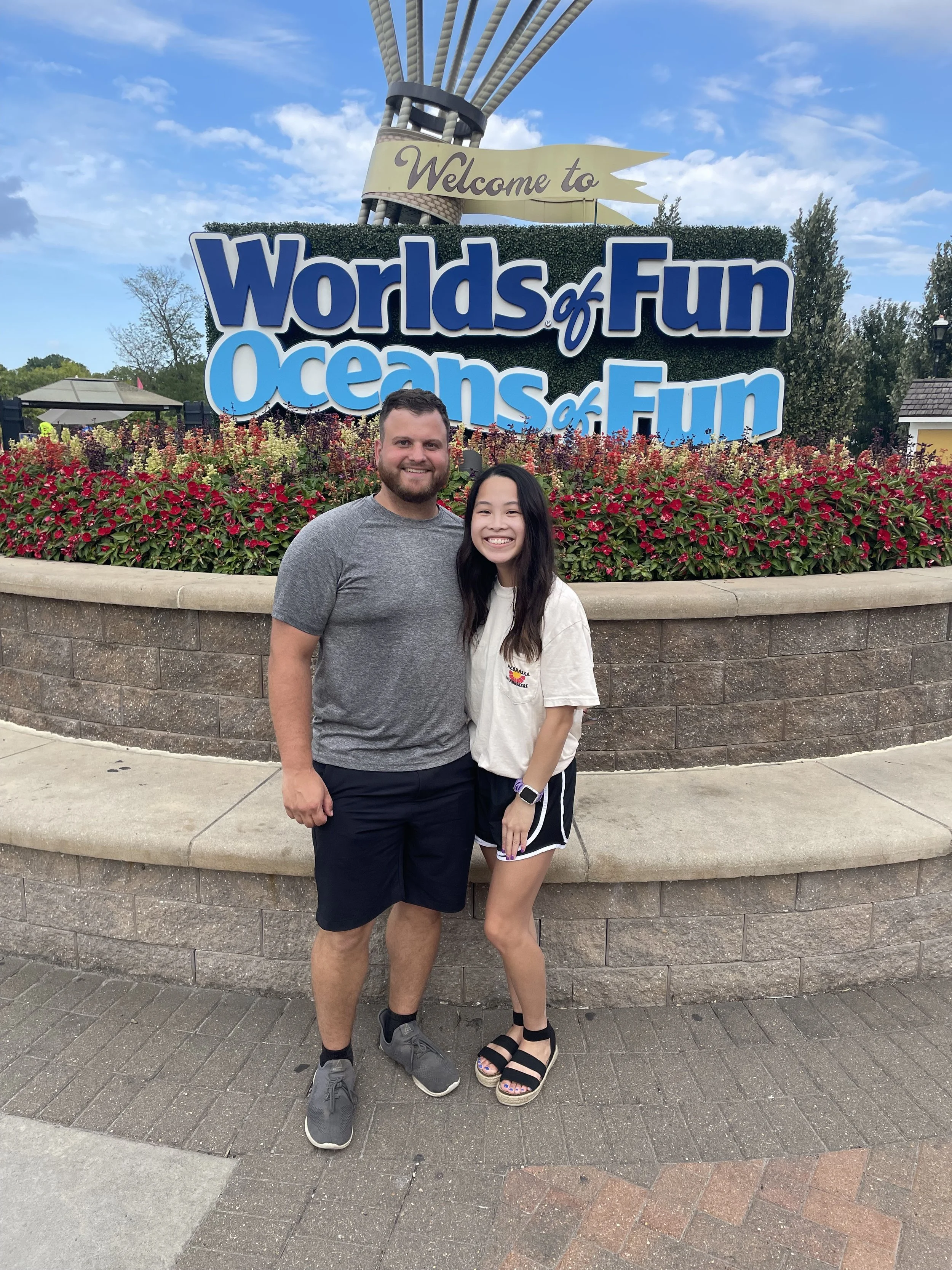 Two people standing in front of a sign that reads 'Welcome to Worlds of Fun Oceans of Fun' with a partly cloudy sky in the background.