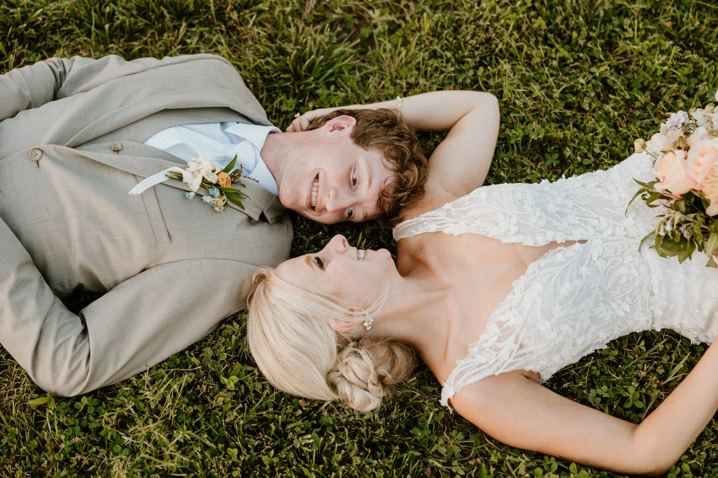 A newlywed couple lies on the grass, looking at each other and smiling, with the bride holding a bouquet of flowers.