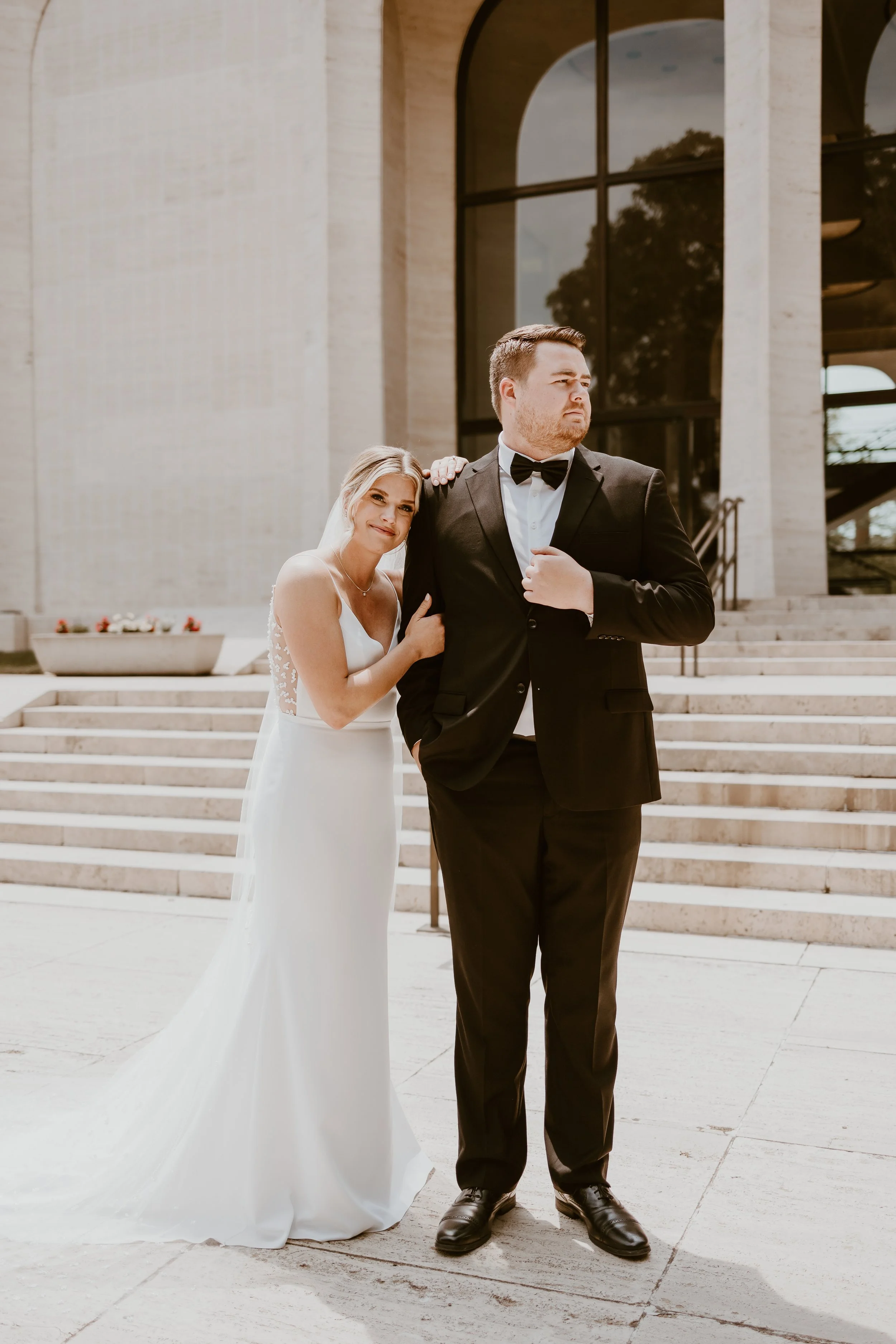 A bride leaning on a groom in a tuxedo outside a modern building with steps and large glass windows.