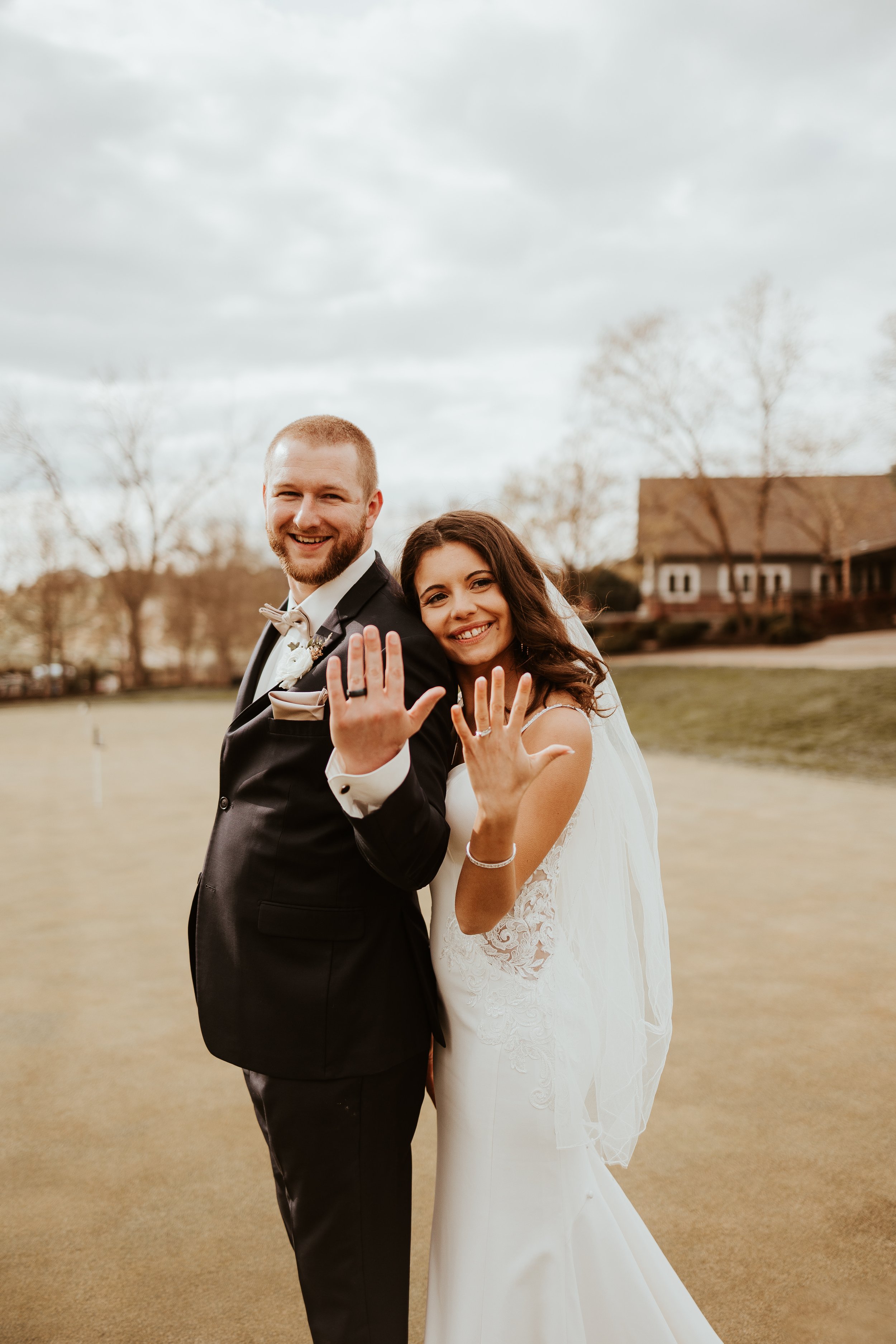 A newlywed couple showing off their wedding rings, smiling and standing on a golf course with trees and a building in the background.