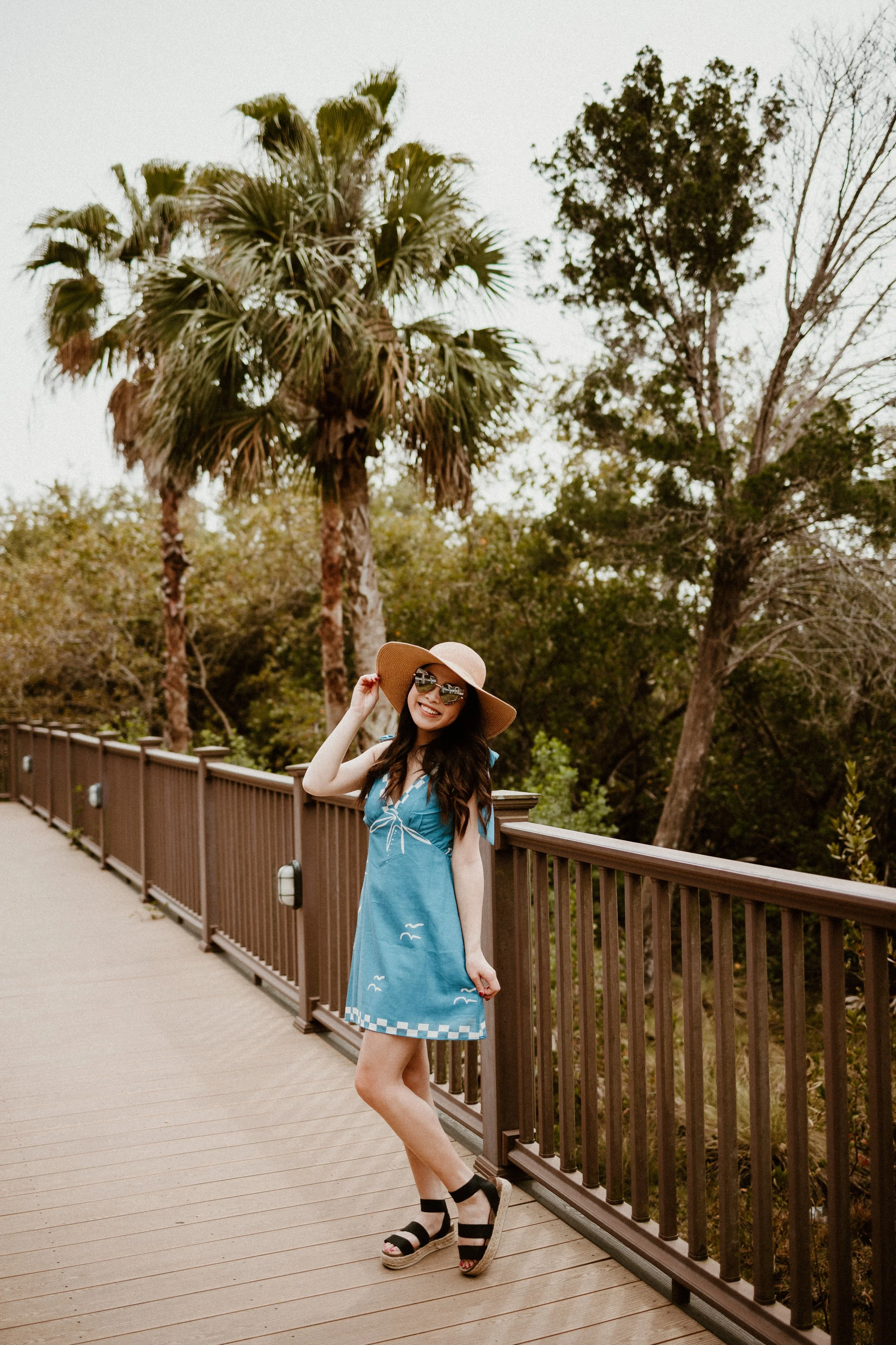 Woman in blue dress and wide-brimmed hat standing on wooden bridge with palm trees and other trees in background, smiling and holding her hat.