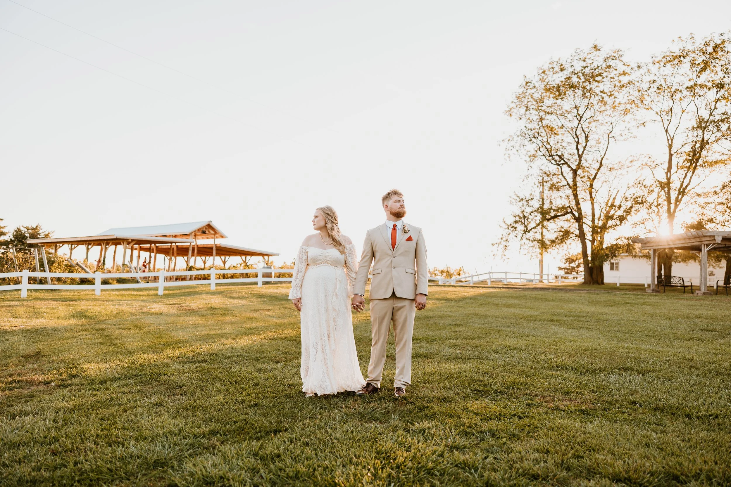 A bride and groom holding hands and standing on a grassy field during sunset, with trees, a white fence, and a pavilion in the background.
