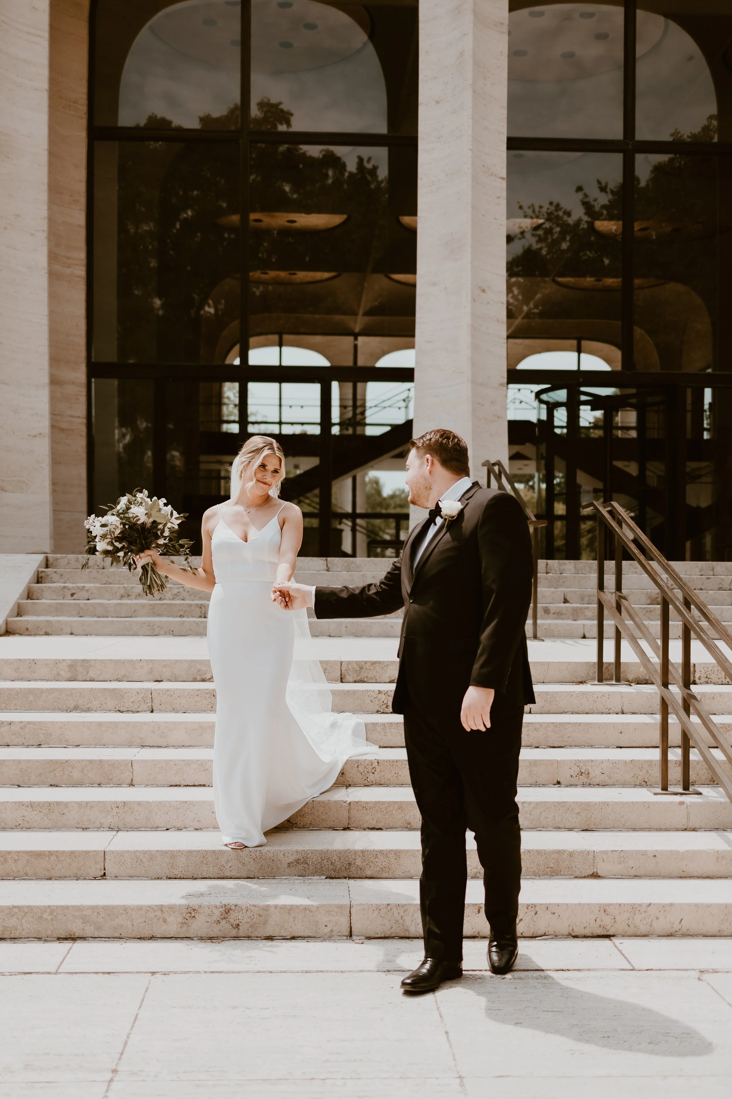 A bride and groom on the stairs outside a modern building, with the bride holding a bouquet and the groom reaching out towards her.