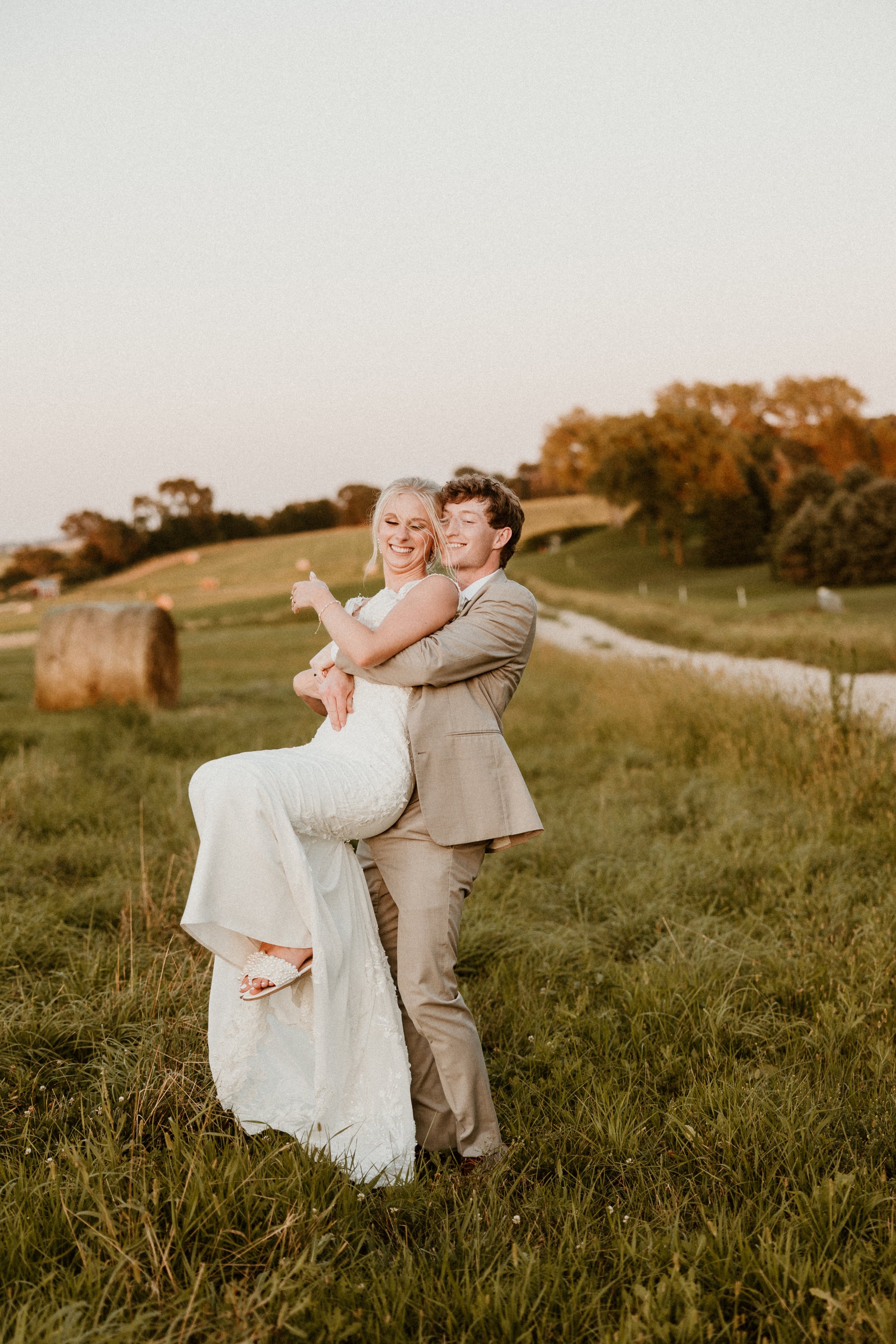 A smiling couple, one in a white wedding dress and the other in a light tan suit, standing outdoors in a grassy field during sunset, with trees and a hay bale in the background.