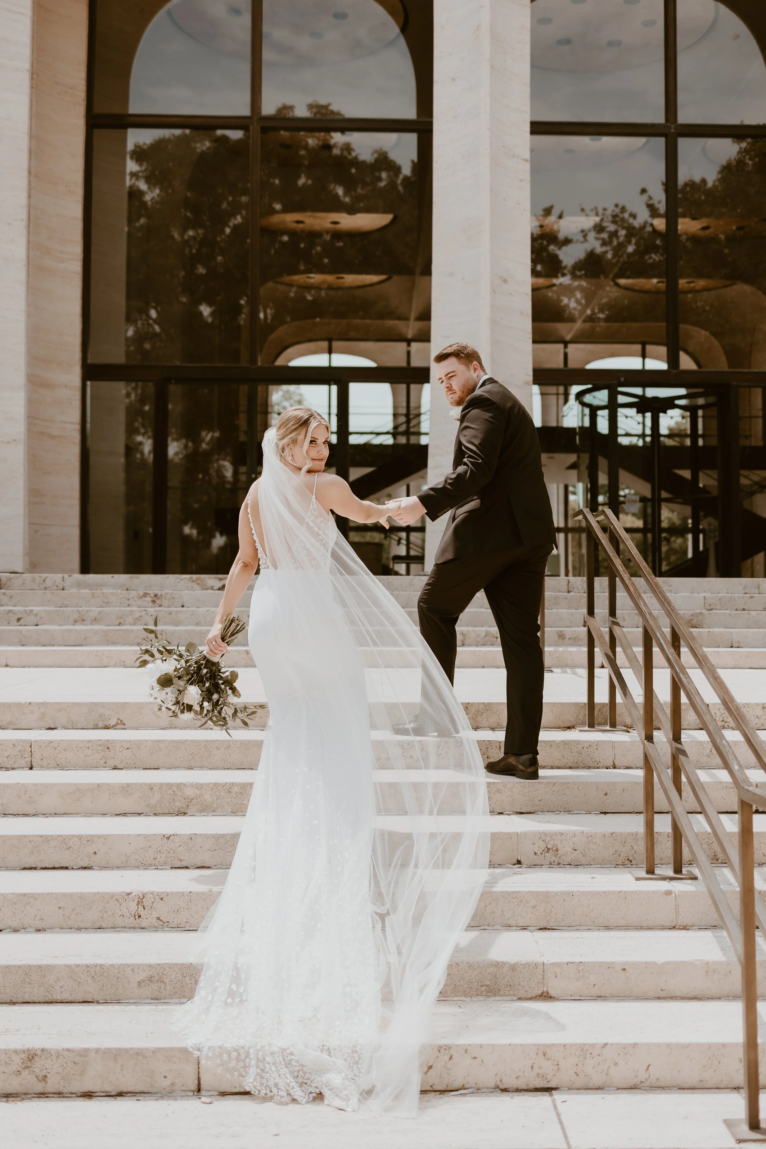 A bride in a white wedding dress and veil holding a bouquet, holding hands with a groom in a black suit and tie, standing on the stairs outside a modern building with large glass windows.