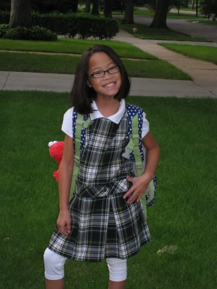 A young girl with glasses, wearing a plaid dress and white leggings, standing on a grassy area with a backpack and smiling.