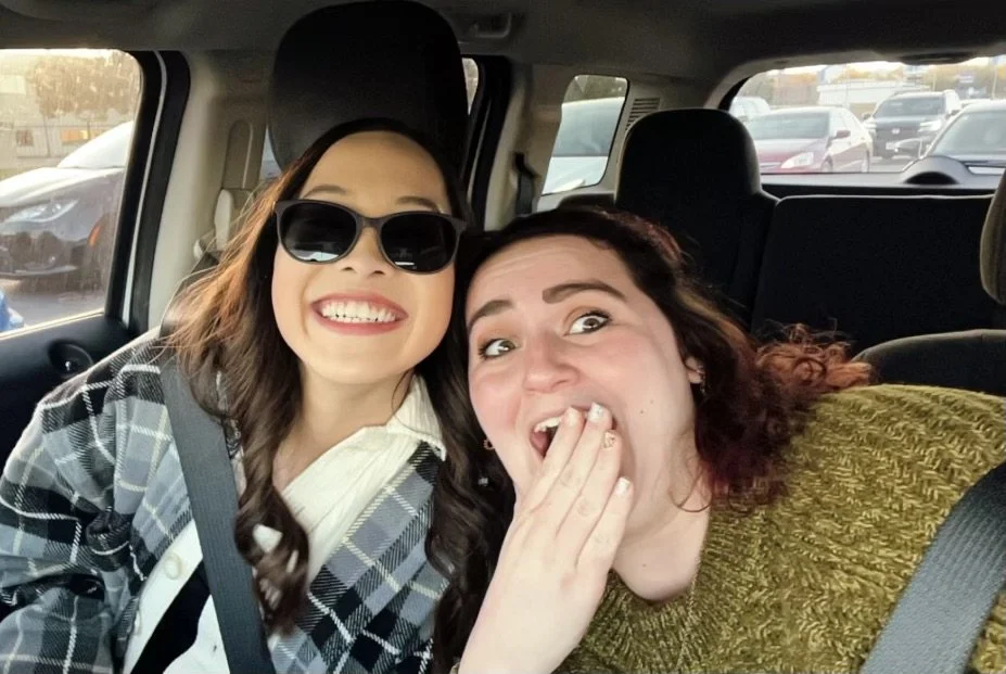 Two women taking a selfie inside a car, smiling and looking cheerful, with traffic visible through the windows.