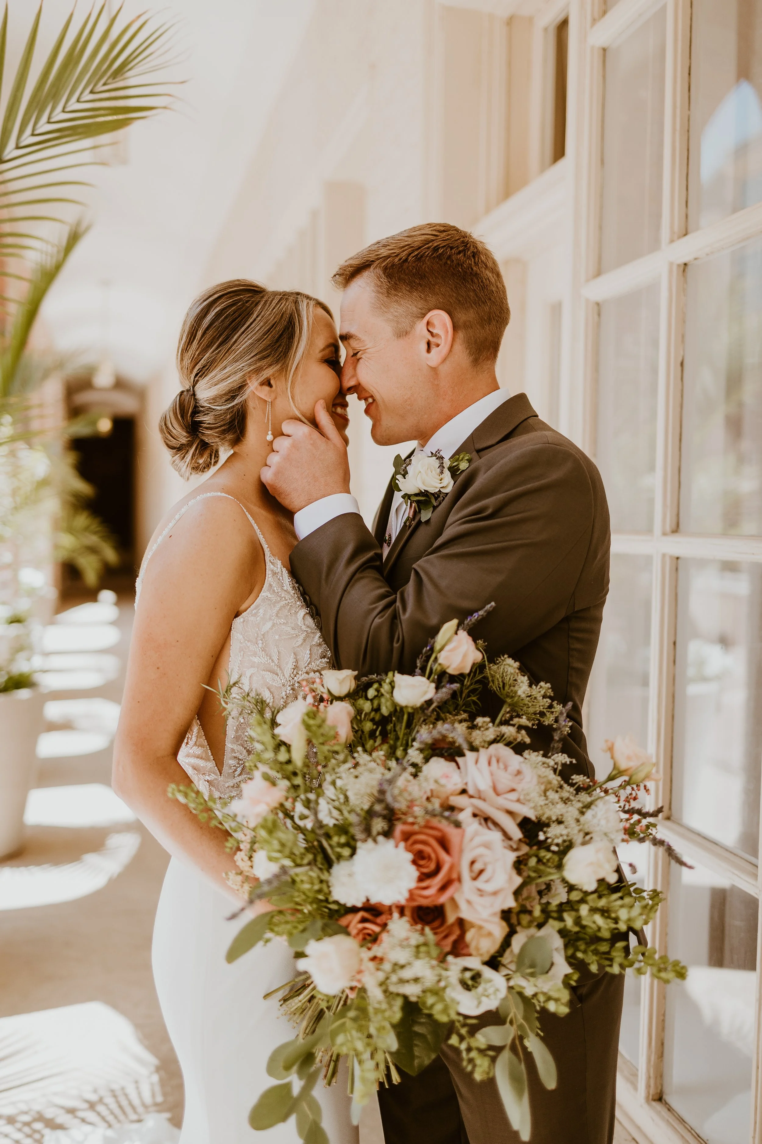 A bride and groom are close together, touching foreheads and smiling, with the groom gently holding the bride's chin. The bride is holding a large bouquet of roses and other flowers. The scene is indoors near a window, with sunlight illuminating the couple.