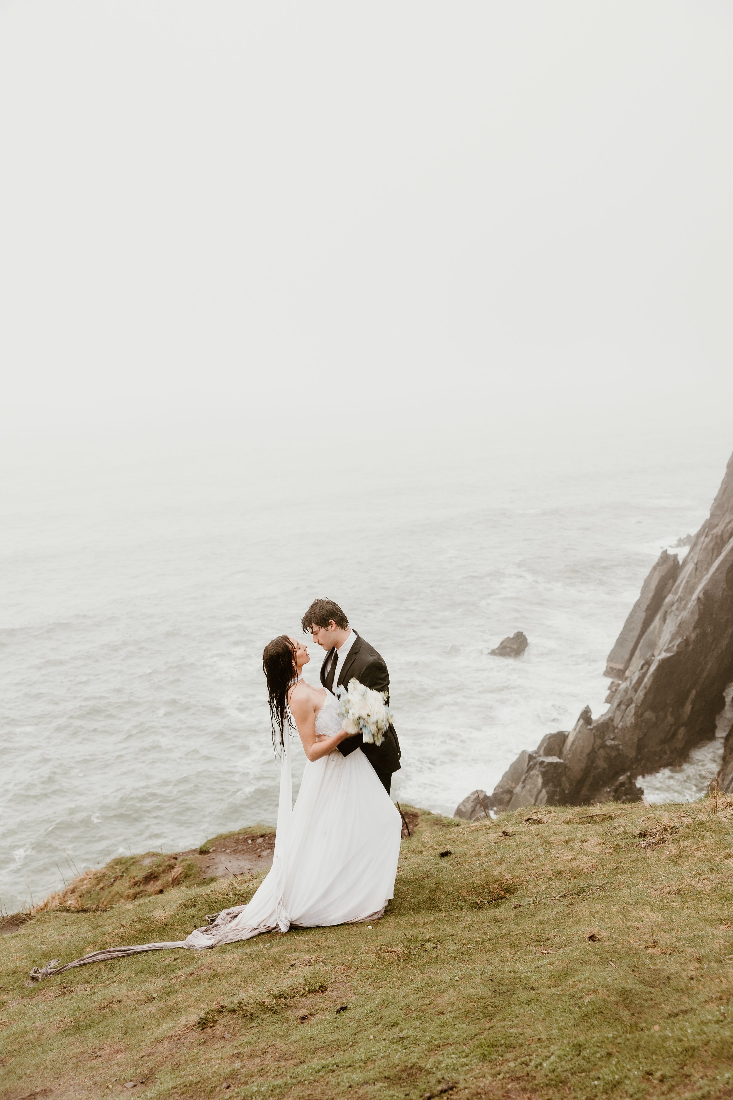 A bride and groom in wedding attire sharing a moment on a grassy cliffside near the ocean, with the bride holding a bouquet of white flowers.
