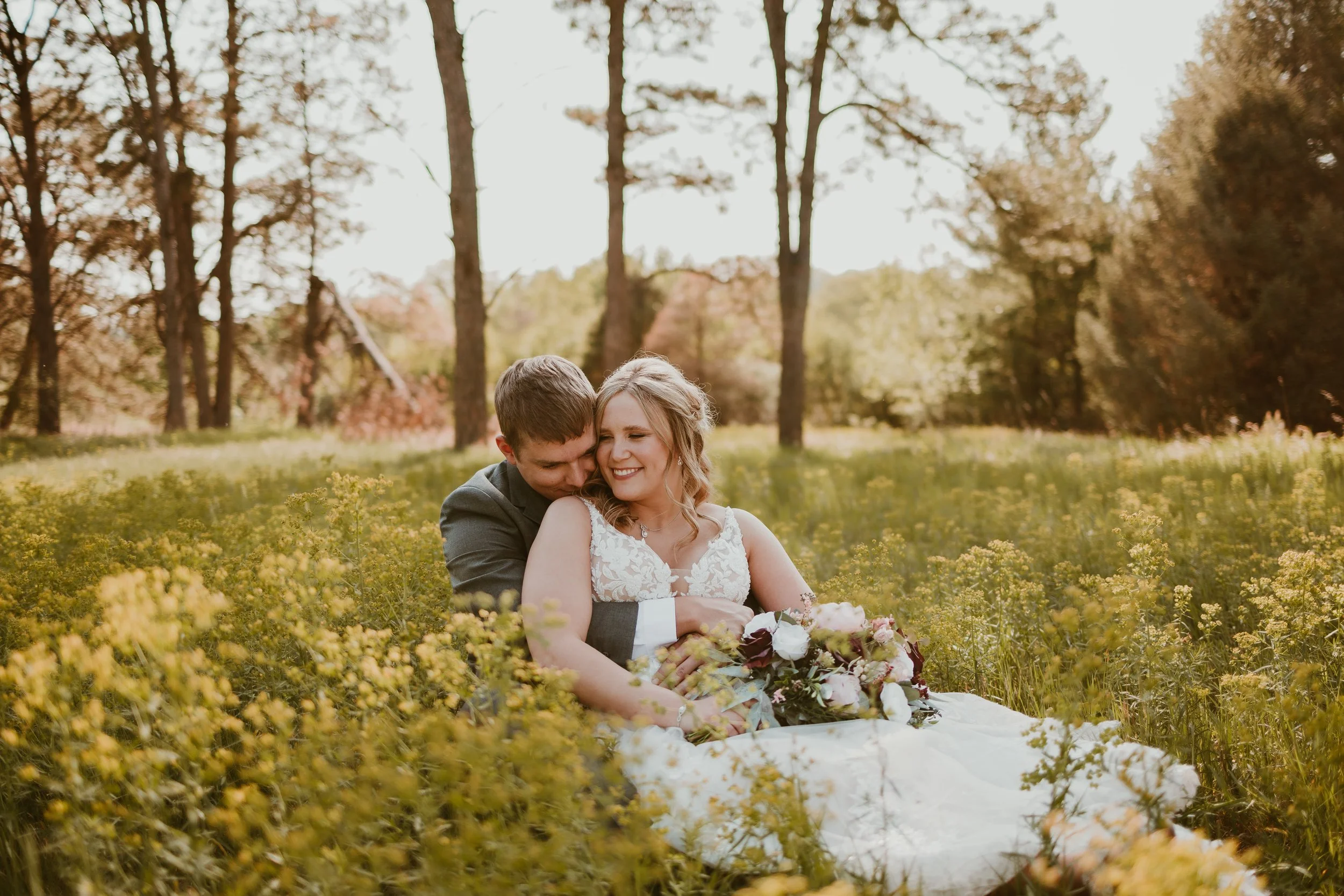 A happy newlywed couple sitting on the grass in a field of yellow flowers, with trees and a clear sky in the background, during sunset.