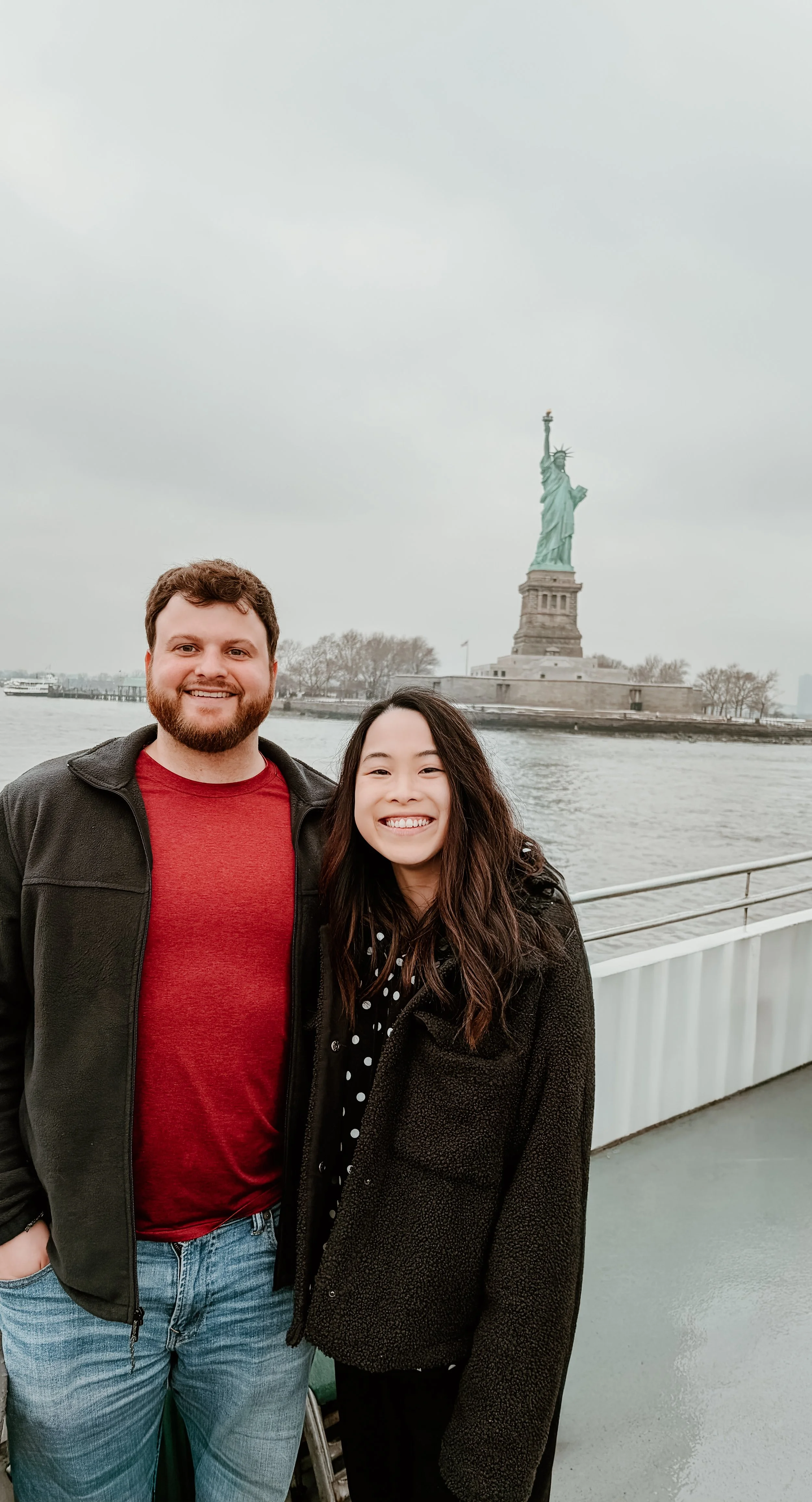 A happy man and woman standing in front of the Statue of Liberty on a cloudy day