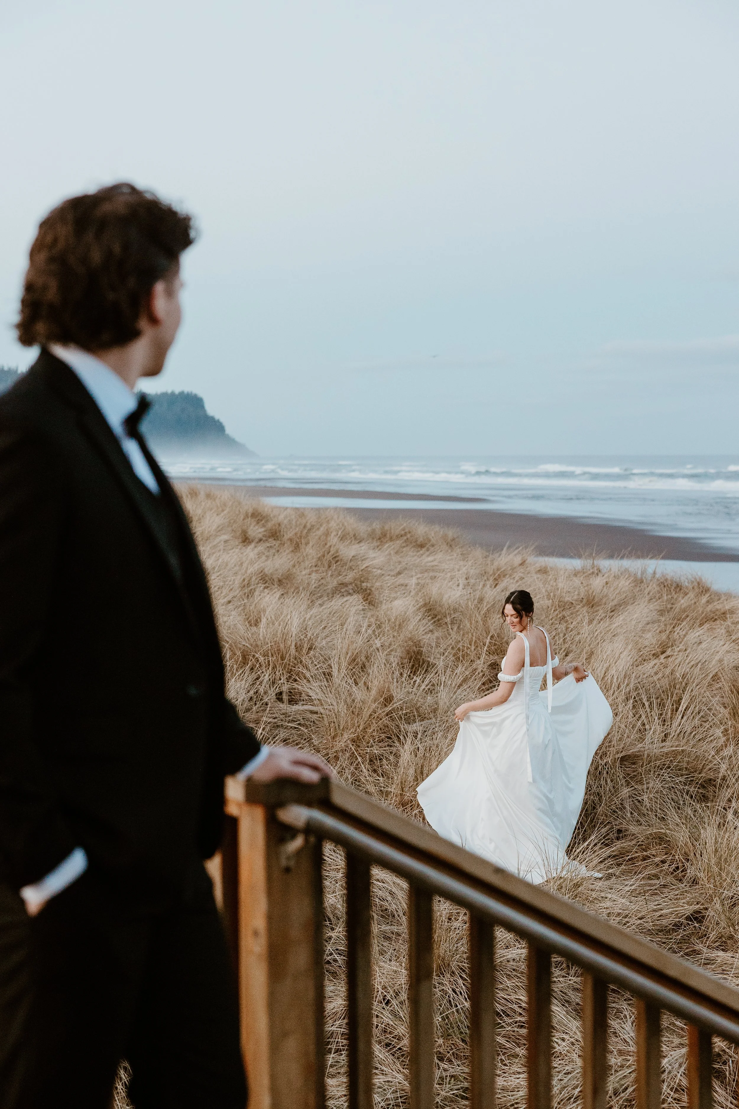 A man in a black tuxedo stands on a wooden staircase, looking towards a woman in a white wedding dress who is walking through tall beach grass with the ocean in the background.