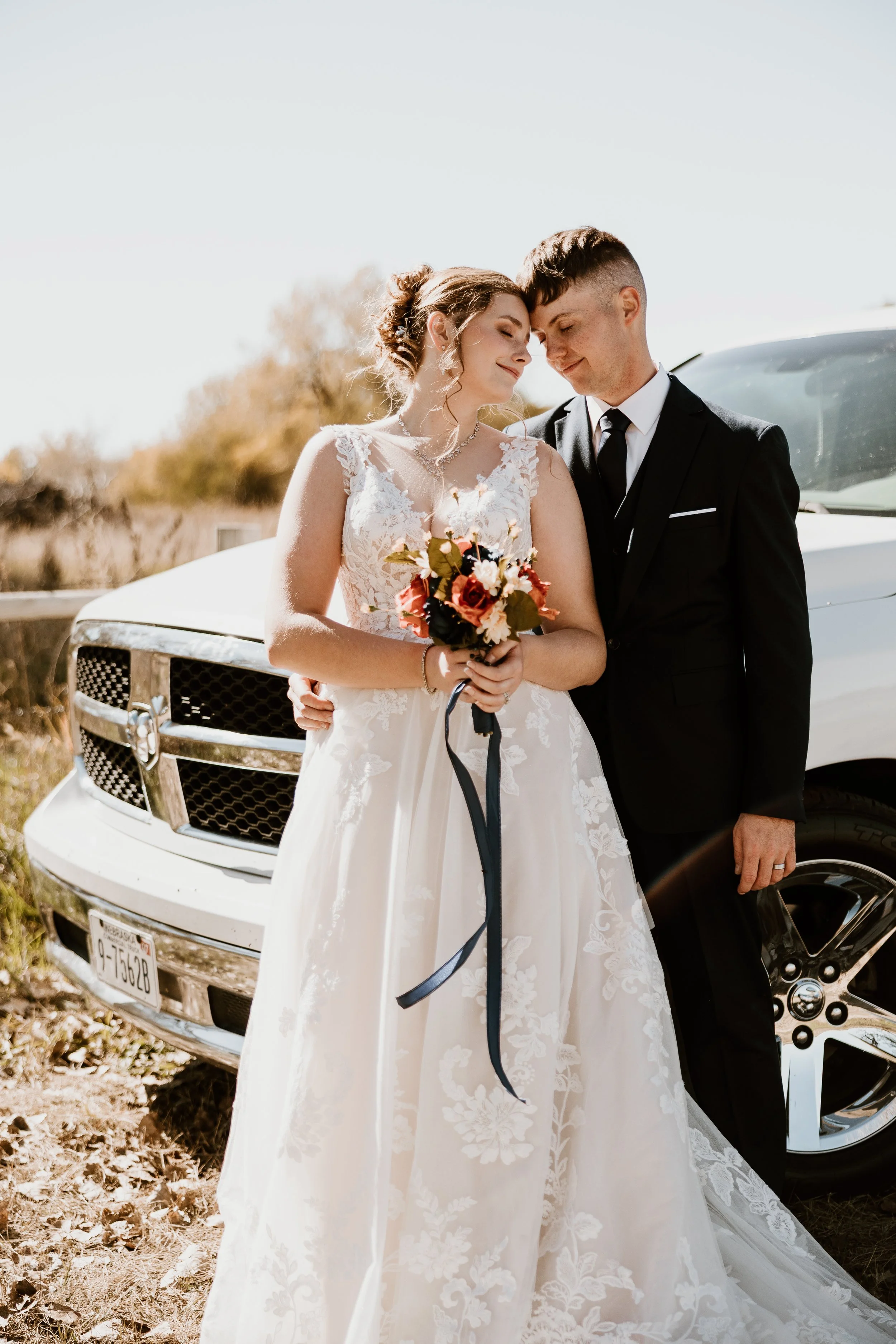 A bride in a white wedding dress and a groom in a black suit stand close together, smiling with foreheads touching, holding a bouquet of flowers, next to a white car outdoors.