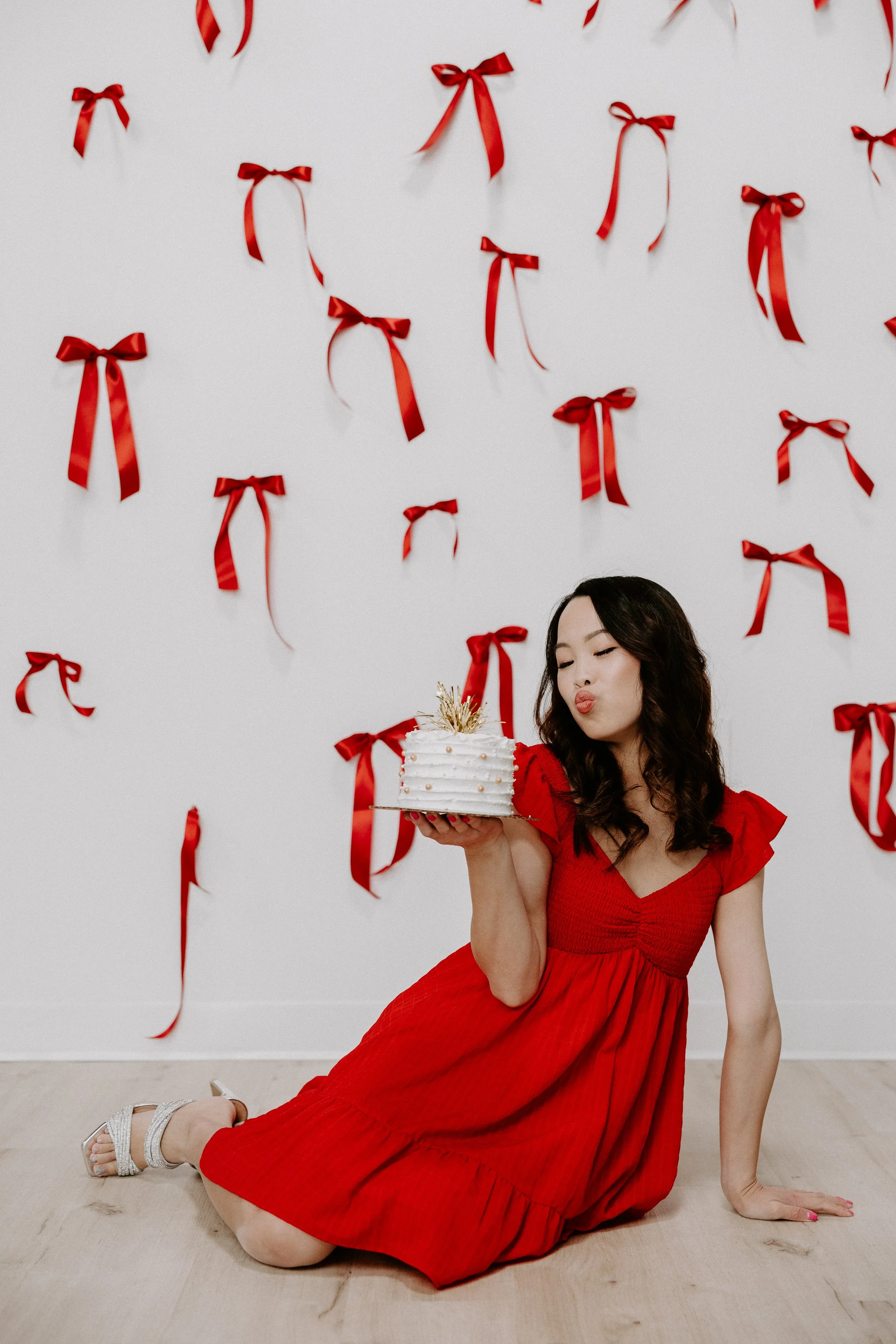 A woman in a red dress is sitting on the floor, holding a cake with white frosting and gold decorations, in front of a wall decorated with red ribbons.