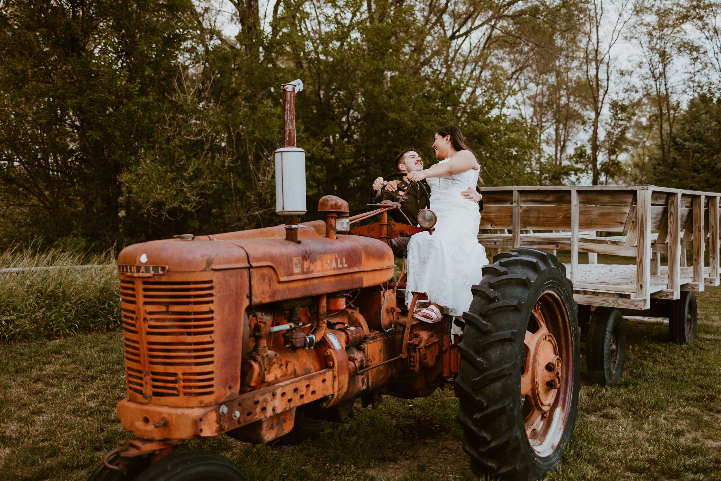 A bride and groom on an old rusty tractor, with the bride sitting on the tractor's hood and the groom standing next to her, outdoors in a grassy field with trees in the background.