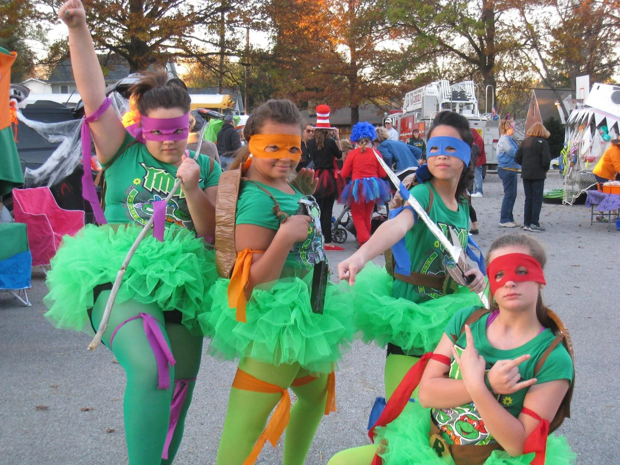 Four young girls in colorful superhero costumes, wearing green tutus, brightly colored tights, and eye masks, posing at an outdoor event or festival.