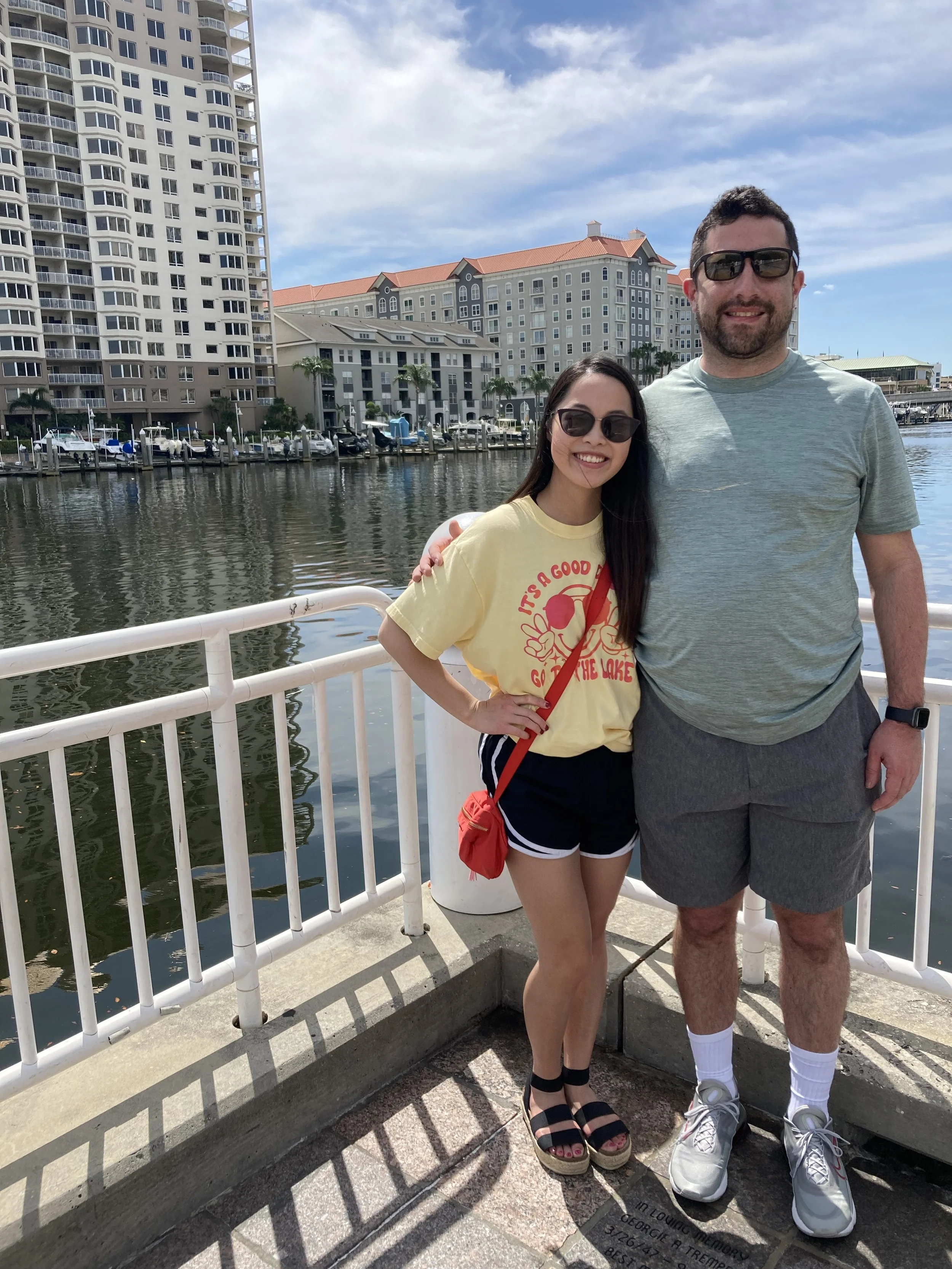 Two people, a woman and a man, stand together on a waterfront promenade in a city with high-rise buildings and a marina in the background. They are smiling, wearing sunglasses, and casual summer clothing.
