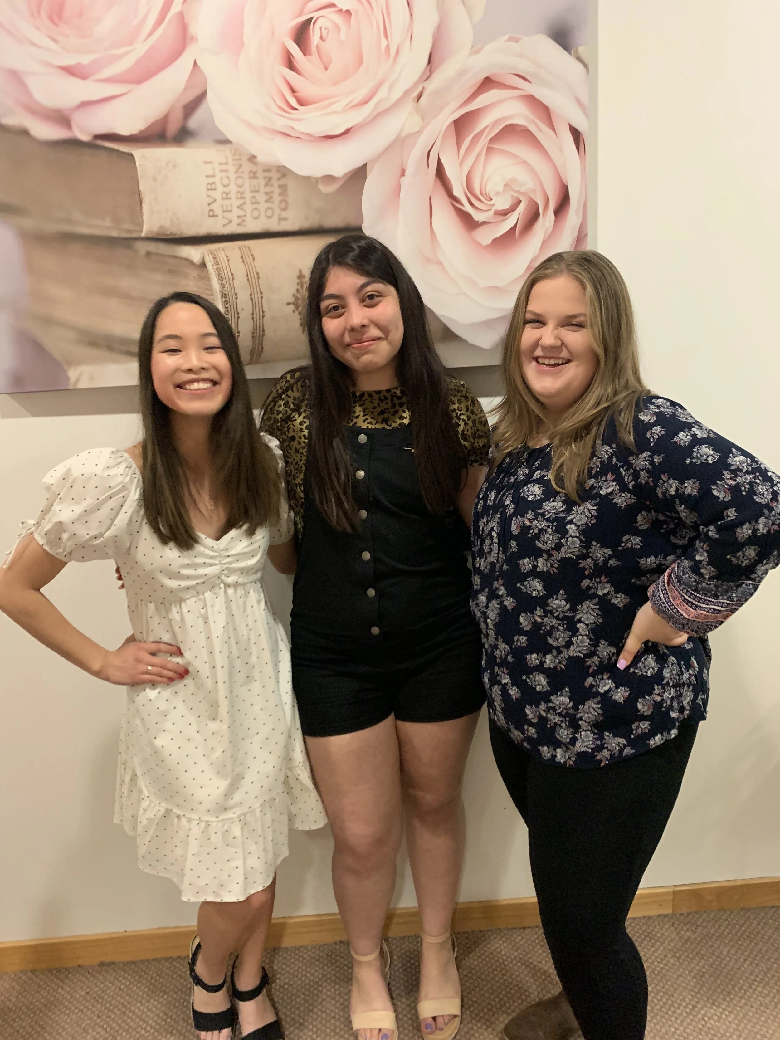 Three women standing together indoors in front of a large floral artwork with pink roses and a book on a white wall, smiling and posing for a photo.