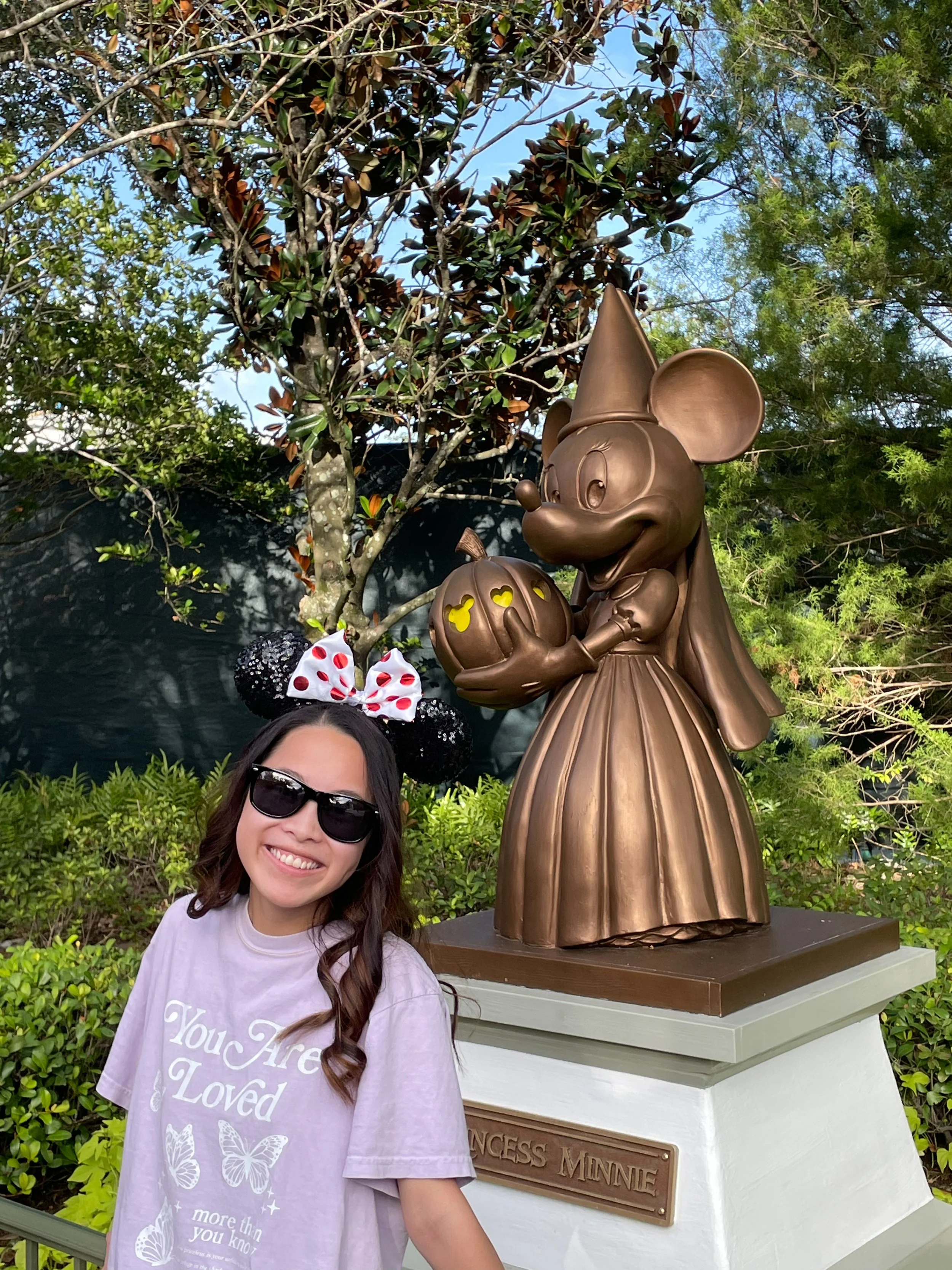 A woman smiling wearing sunglasses and Minnie Mouse ears headband with a white bow with red polka dots, standing in front of a bronze statue of Minnie Mouse holding a pumpkin, outdoors with trees and shrubbery in the background.
