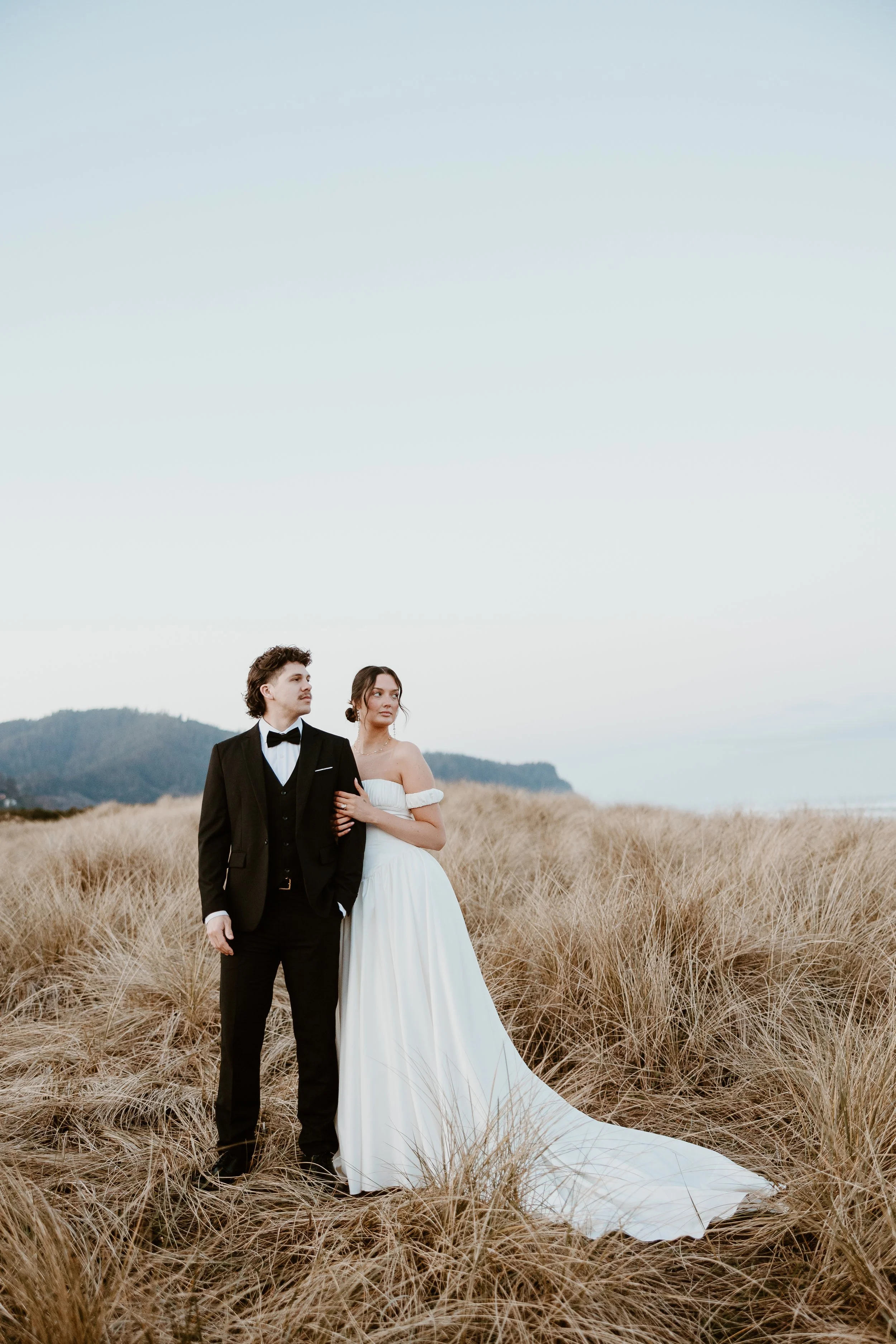 A couple dressed in wedding attire standing in a field of tall grass with a backdrop of hills and a clear sky.