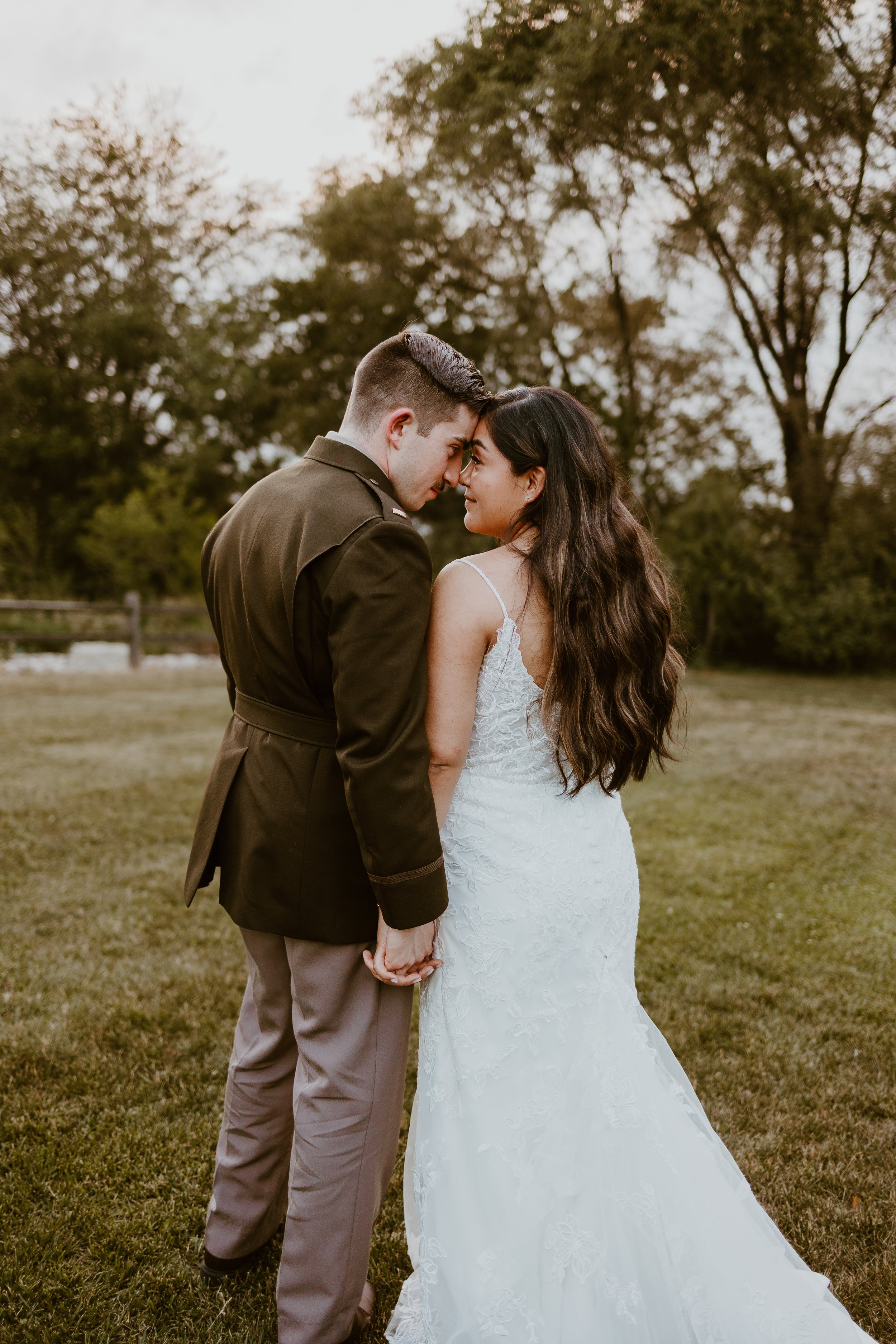 A couple in wedding attire holding hands, standing close with foreheads touching outdoors on a grassy field with trees in the background.