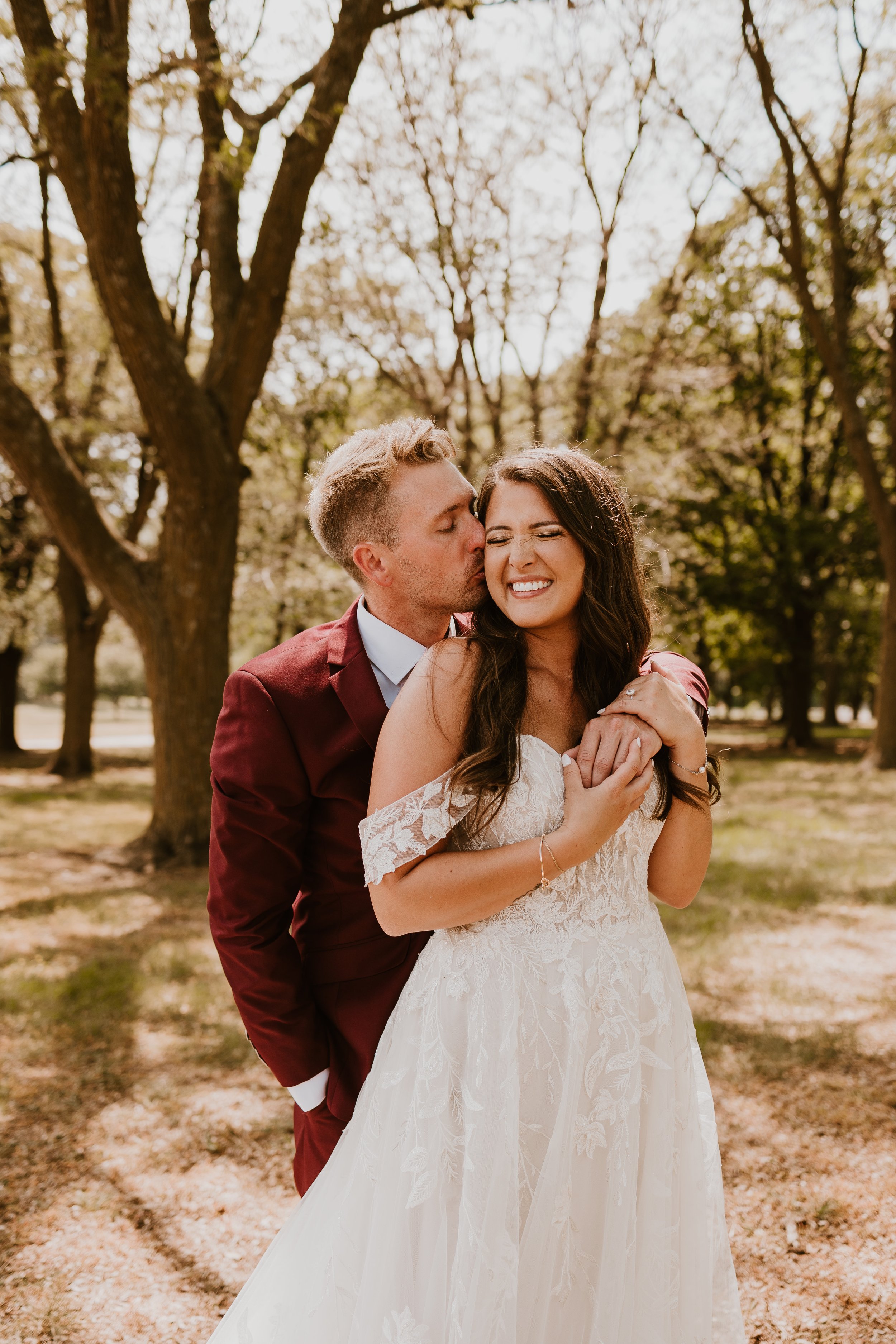 A couple in wedding attire stands outdoors among trees. The man leans in to kiss the woman on the cheek, and she is smiling with her eyes closed while holding his hands around her shoulders.