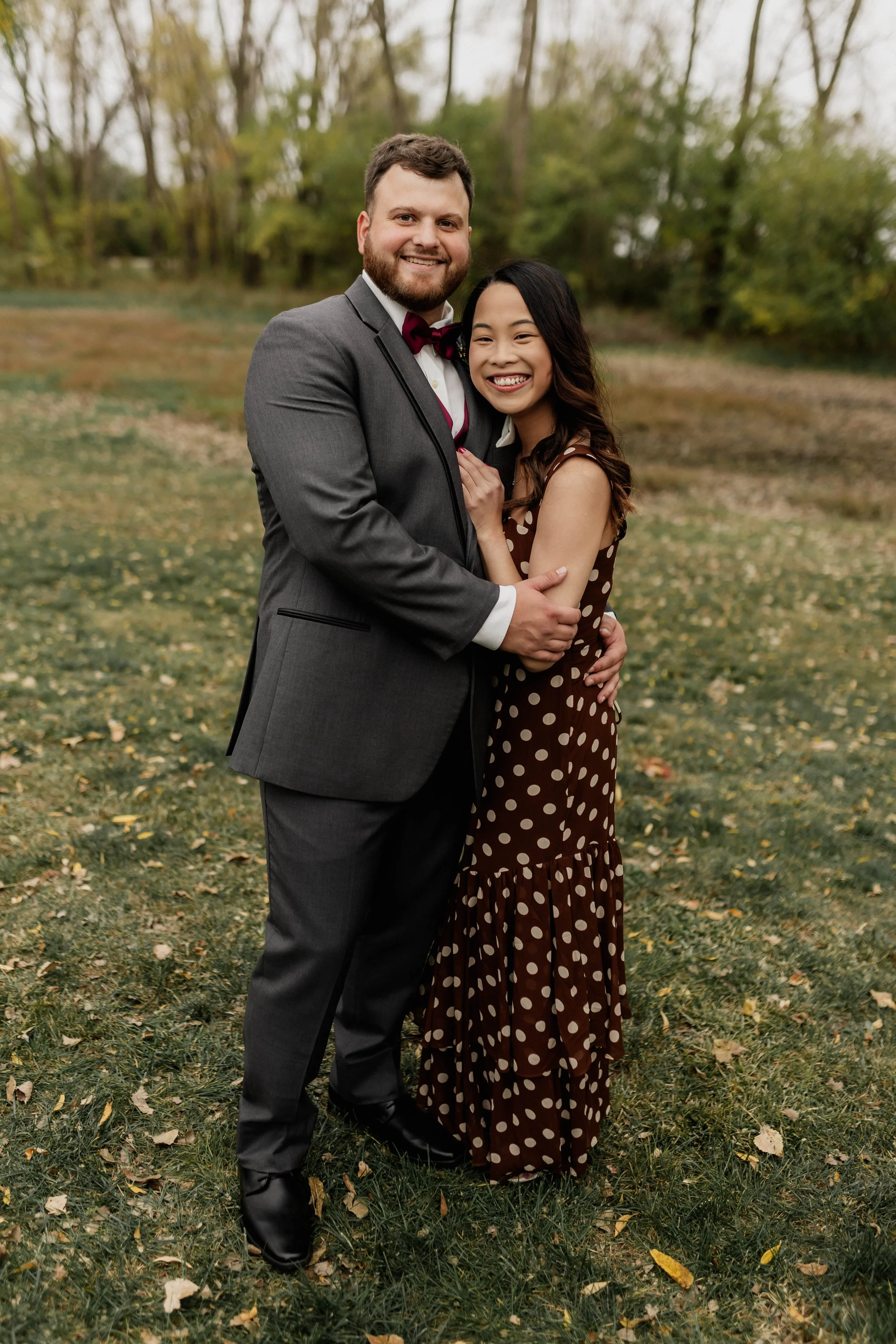 A man and woman standing outdoors, smiling and embracing, with trees and fallen leaves in the background.