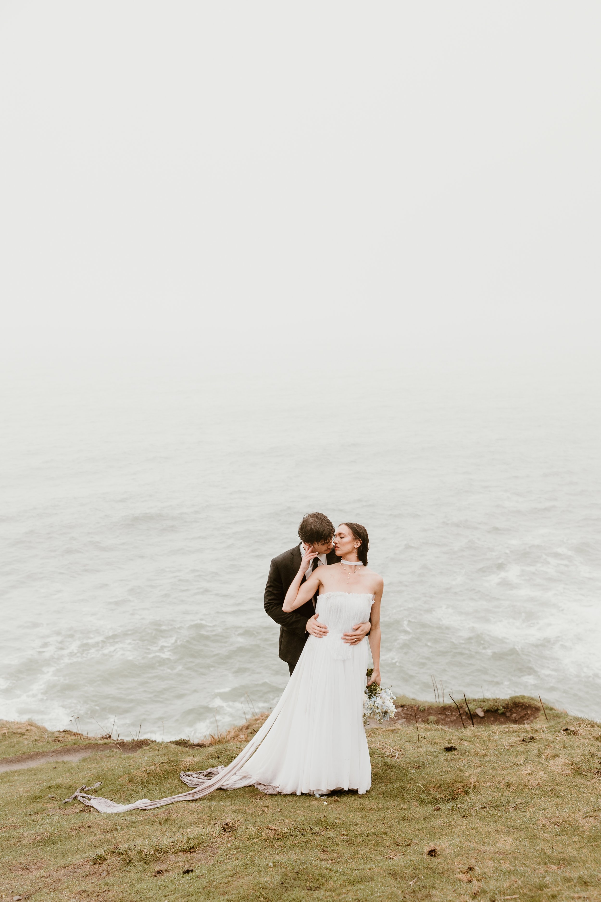 A bride and groom embrace on a grassy cliff overlooking the ocean, with a misty horizon in the background.