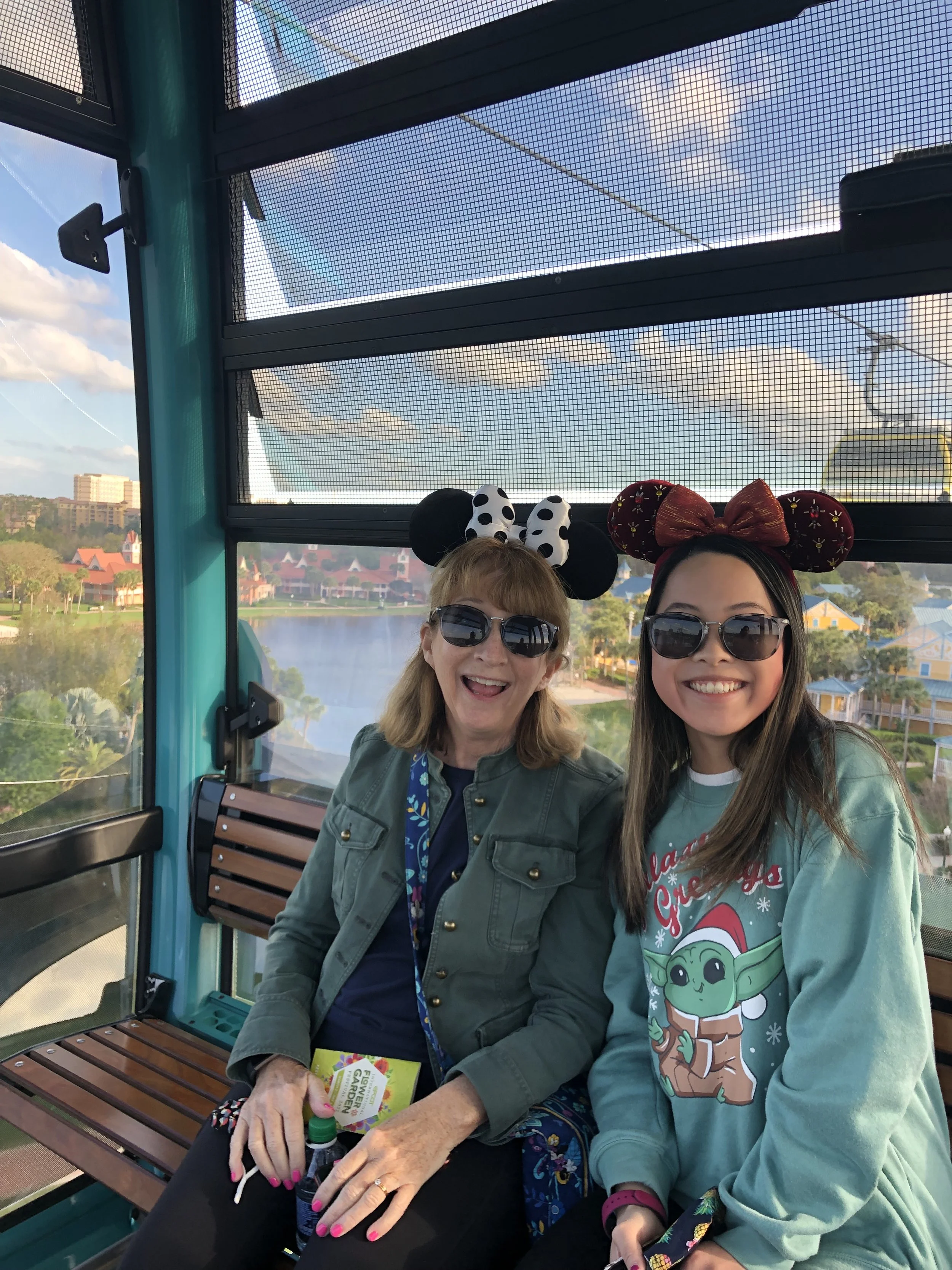 Two women smiling and wearing Minnie Mouse and Christmas-themed Mickey Mouse ears, sunglasses, sitting inside a Ferris wheel cabin with a view of a lake and colorful buildings outside.