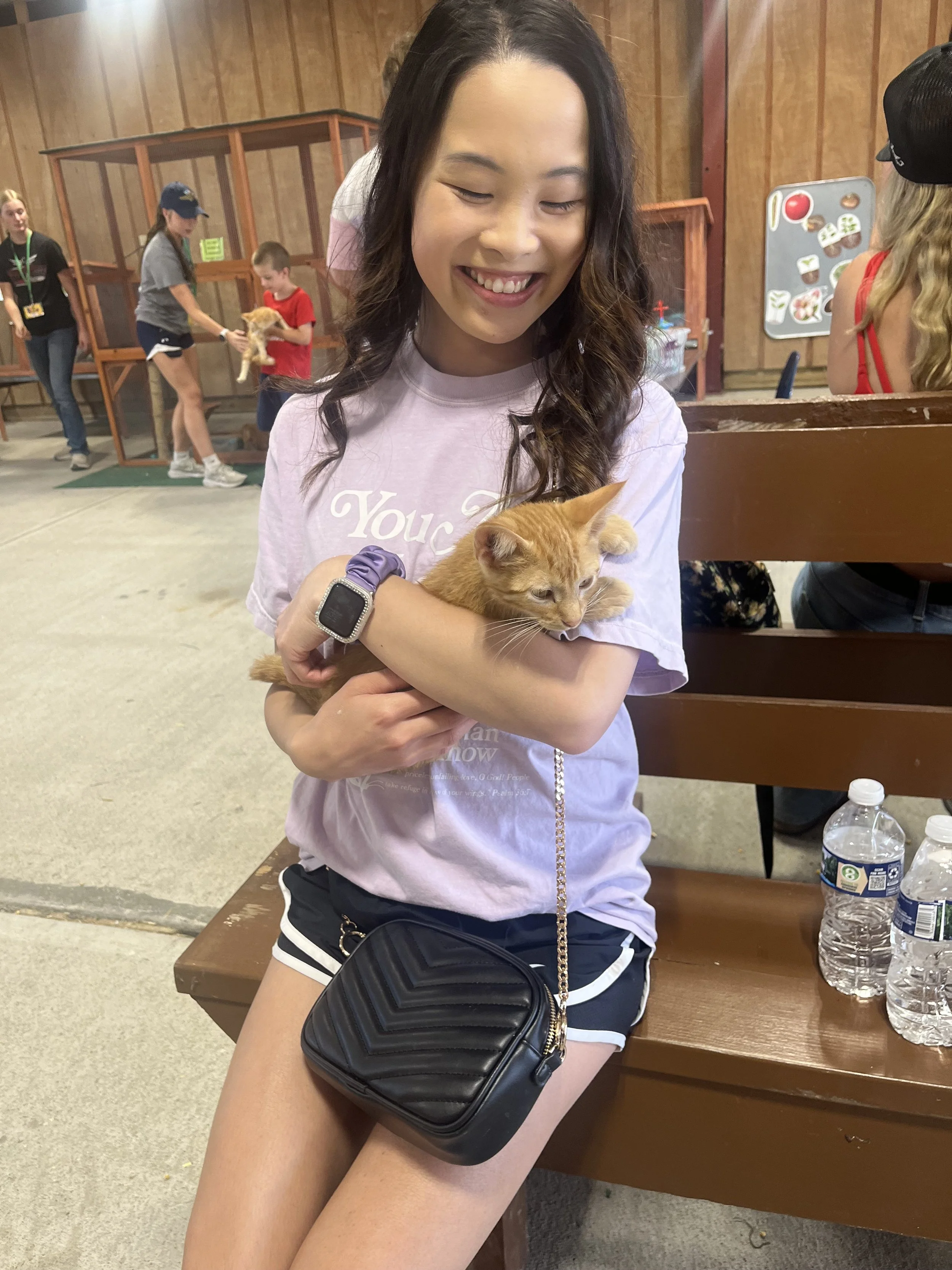 A young woman with long dark hair smiling while holding an orange kitten in her arms, sitting on a wooden bench in a room with wooden walls, with other people and animals in the background.