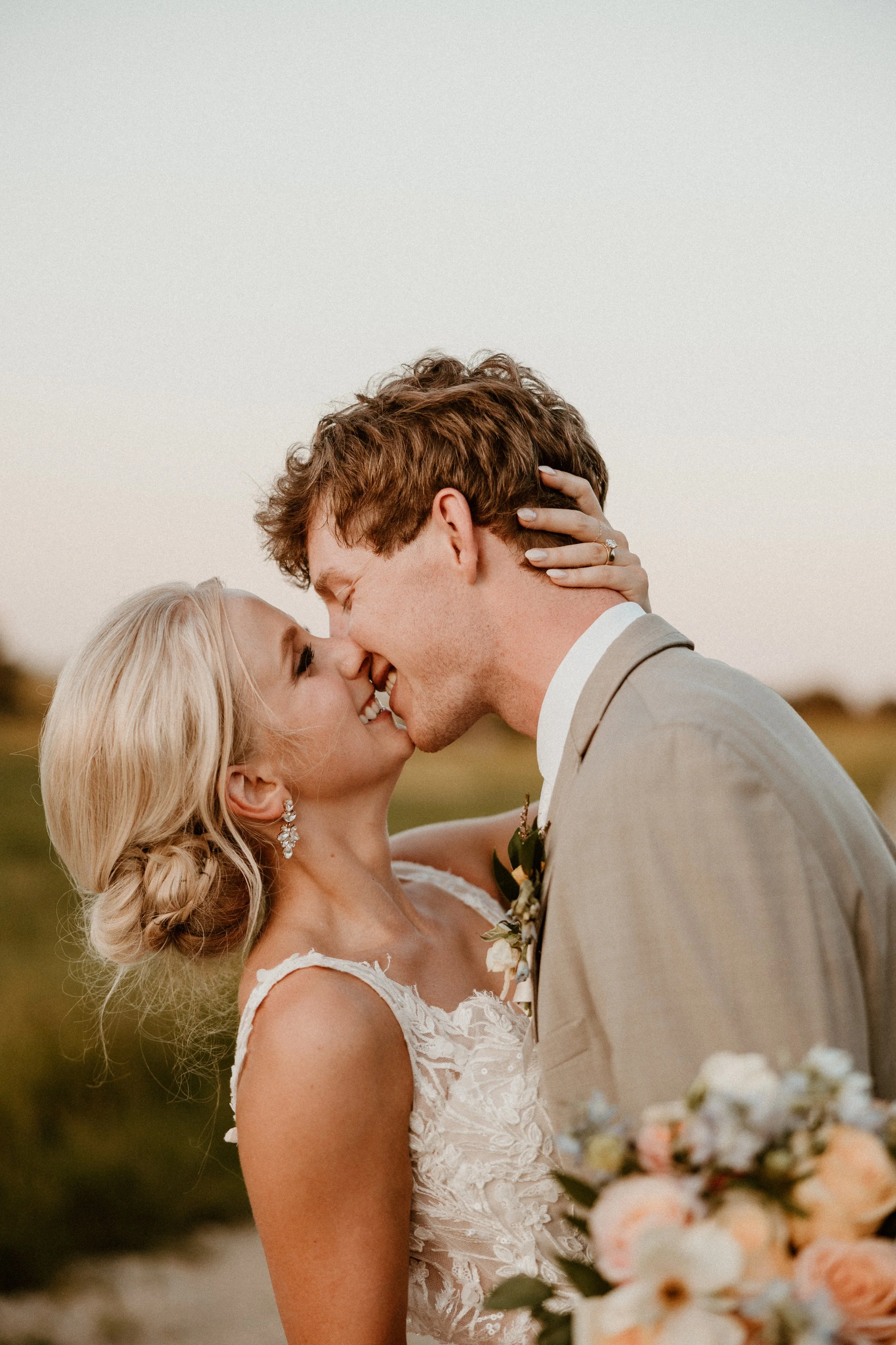 A bride and groom are close together, smiling and about to kiss outdoors at sunset.