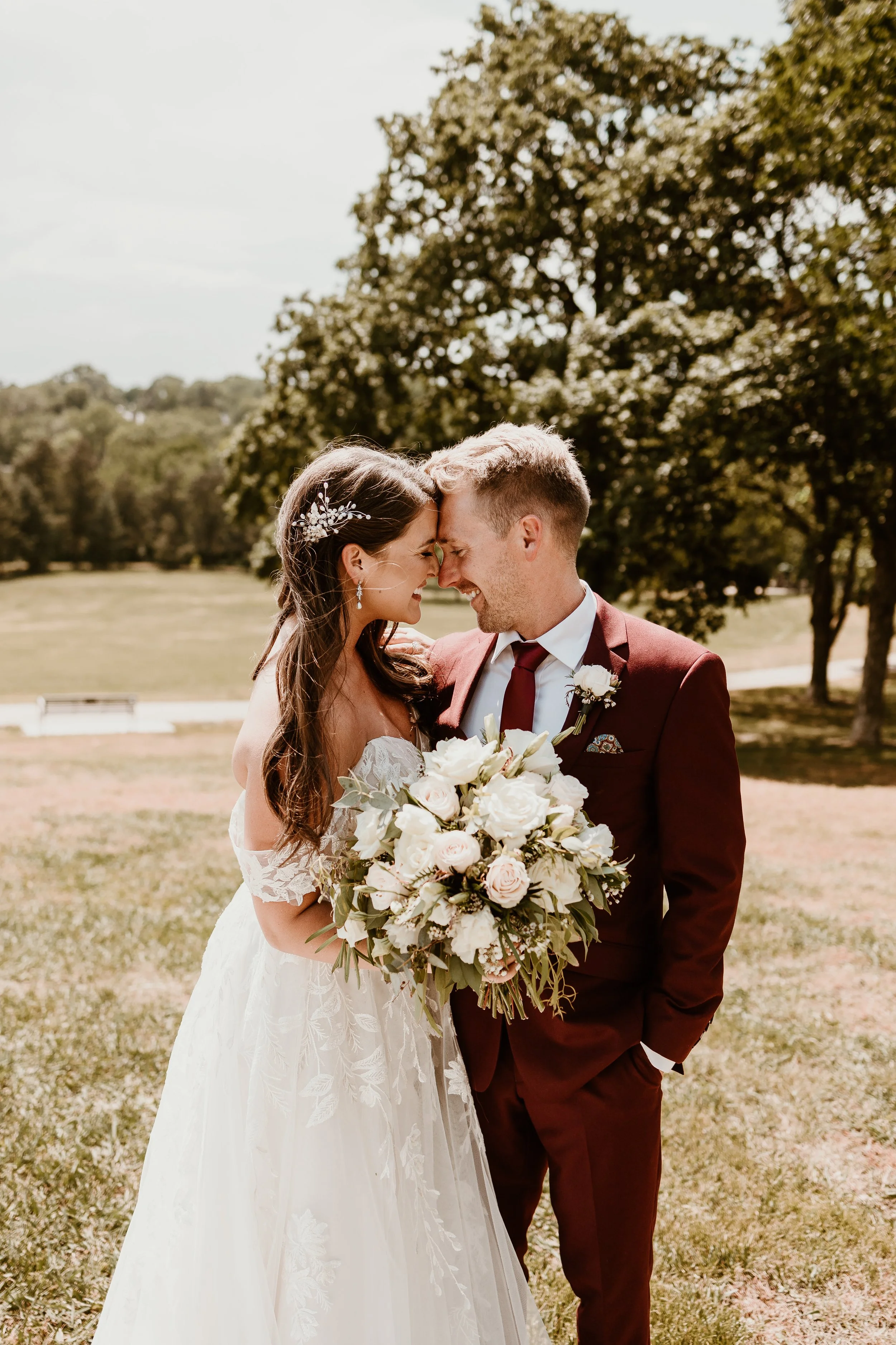 A bride and groom share a joyful moment outside on a sunny day, with their foreheads touching and eyes closed. The bride holds a large bouquet and wears a white wedding dress with lace details. The groom wears a maroon suit and tie, with a boutonnière.