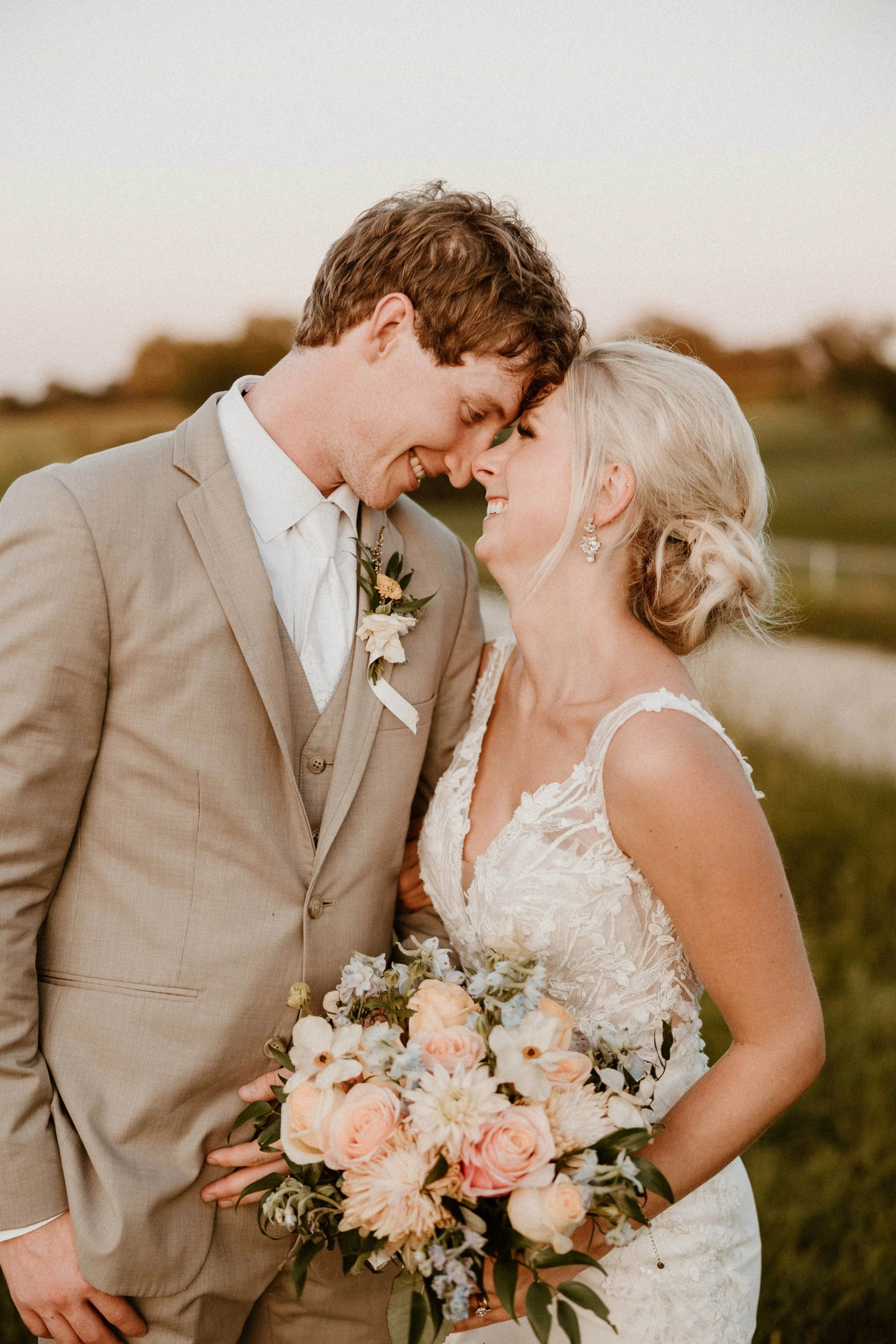 A newlywed couple shares a tender moment outdoors, with foreheads touching, holding a bouquet of blush roses, peonies, and other flowers, dressed in wedding attire, surrounded by a natural landscape.