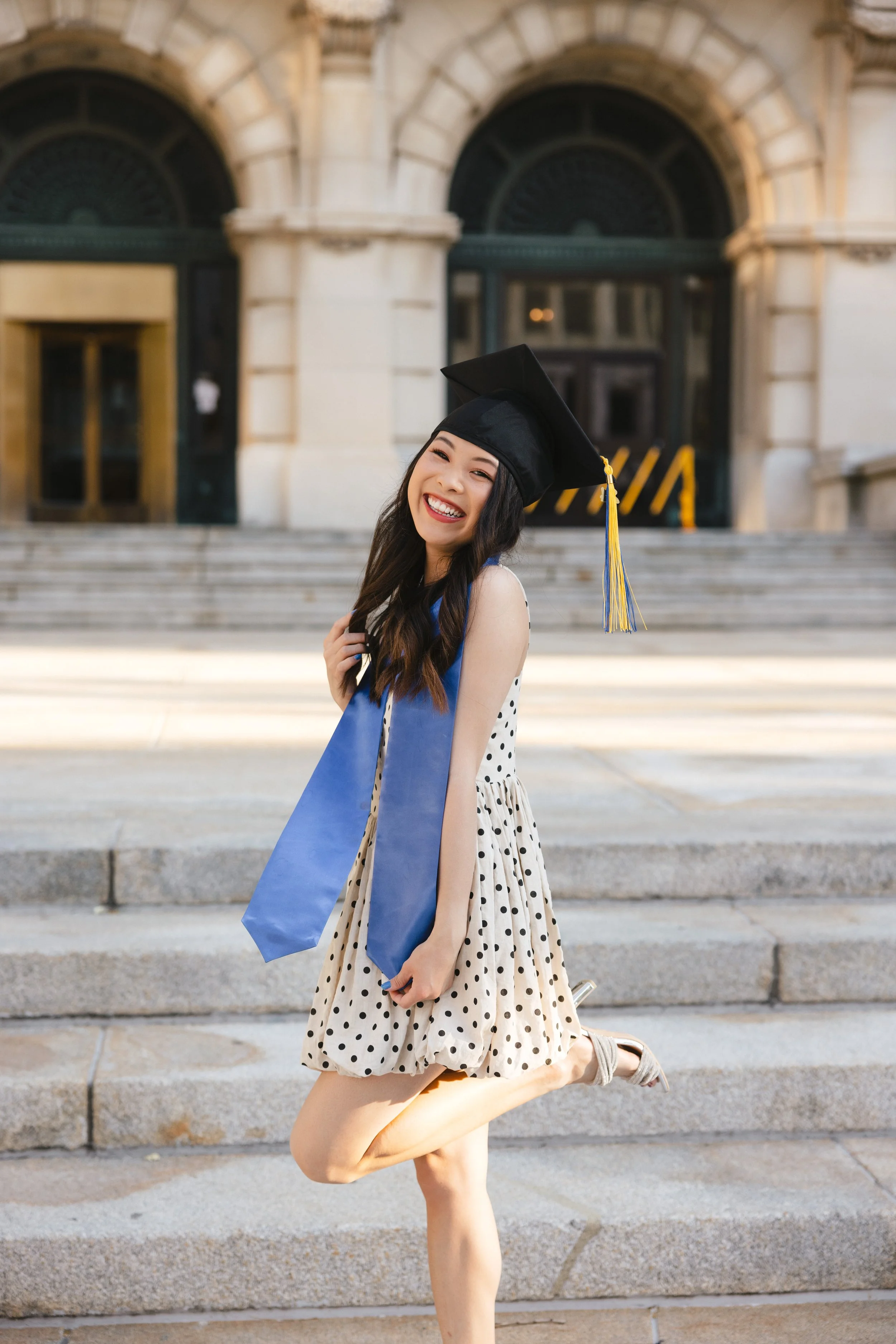 Eager young woman in polka dot dress celebrates graduation, wearing tassel cap and honor stole, smiling with one leg uplifted in front of university steps.
