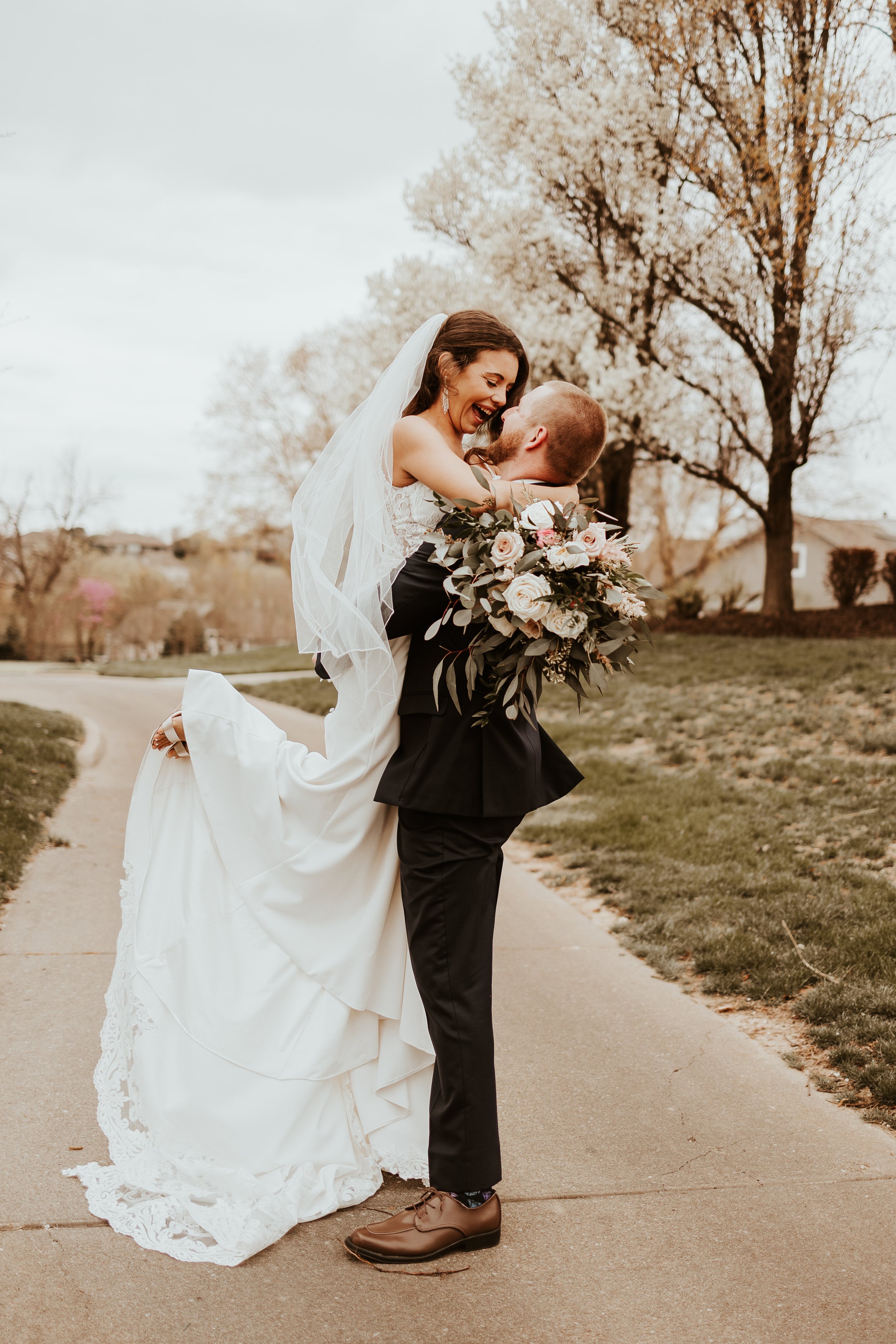 Groom lifting bride in a park with blooming trees and cloudy sky.
