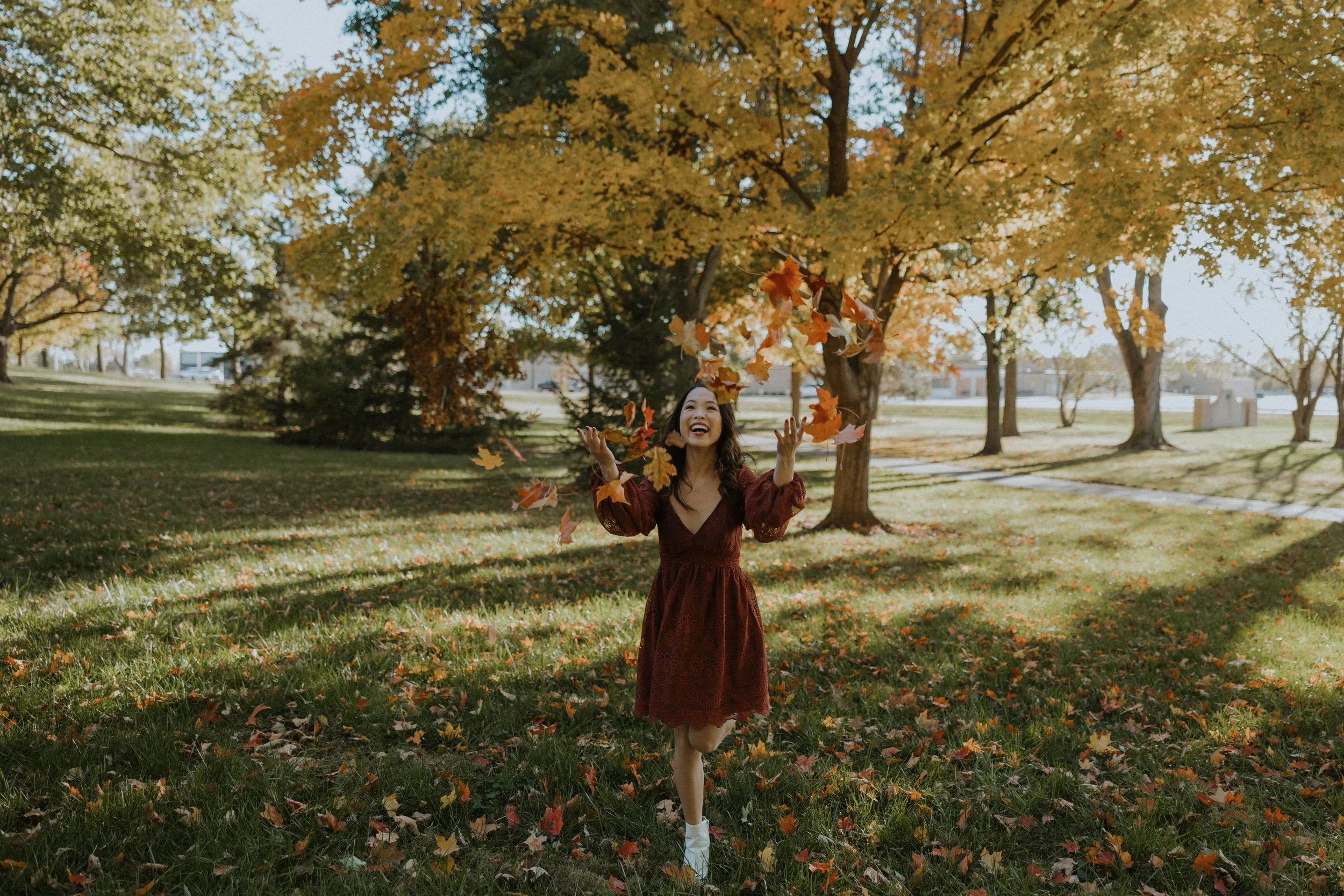 A woman in a burgundy dress enjoying fall leaves in a park with trees and a grassy area.